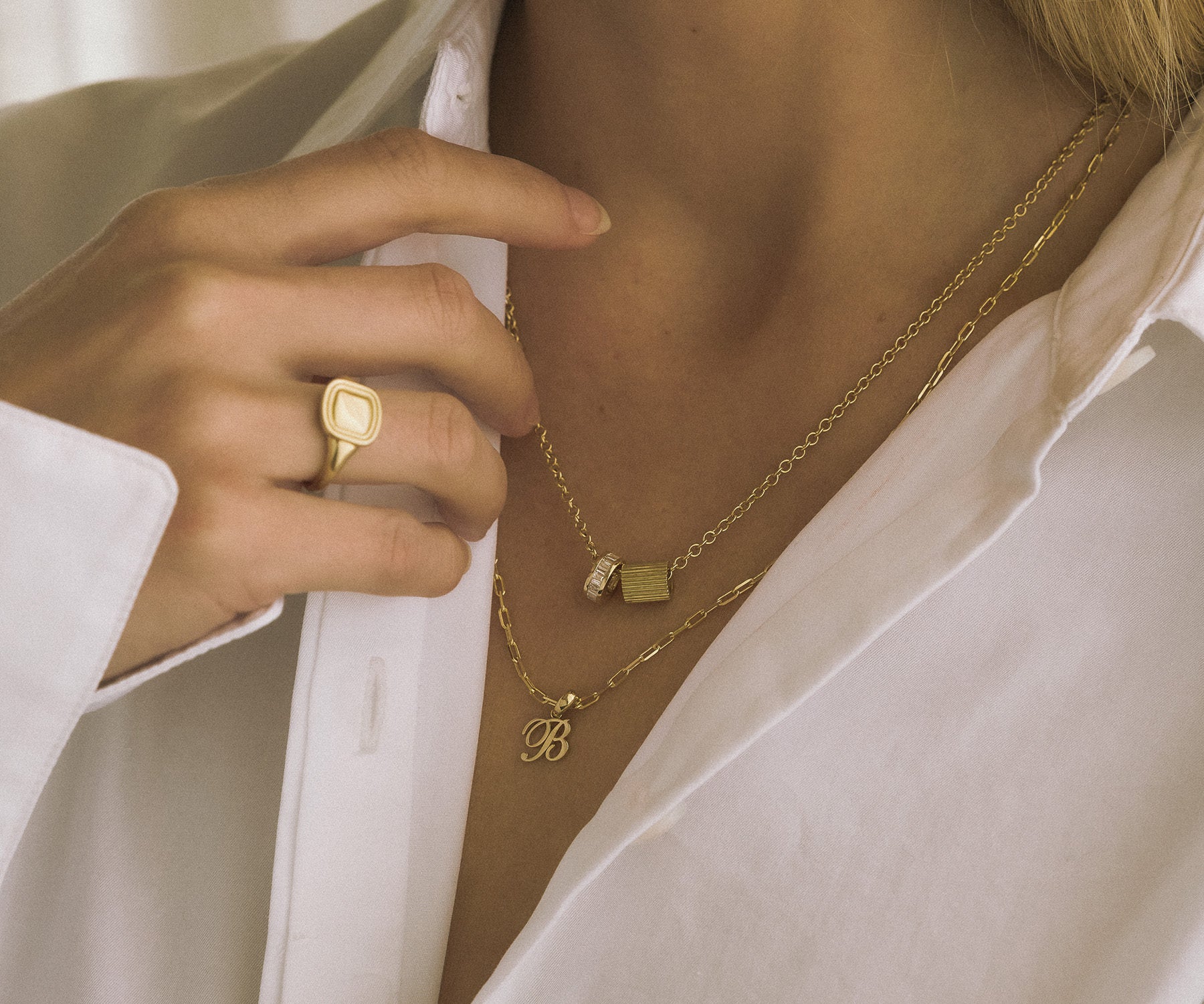Close up of a woman wearing a Kinn x Shoppe Amber Interiors Estate Signet Ring with a Milestone Baguette Diamond Charm and an Estate Charm on a Cable Rolo Chain Necklace layered with a Love Letter Charm Pendant on a Micro Link Chain Necklace