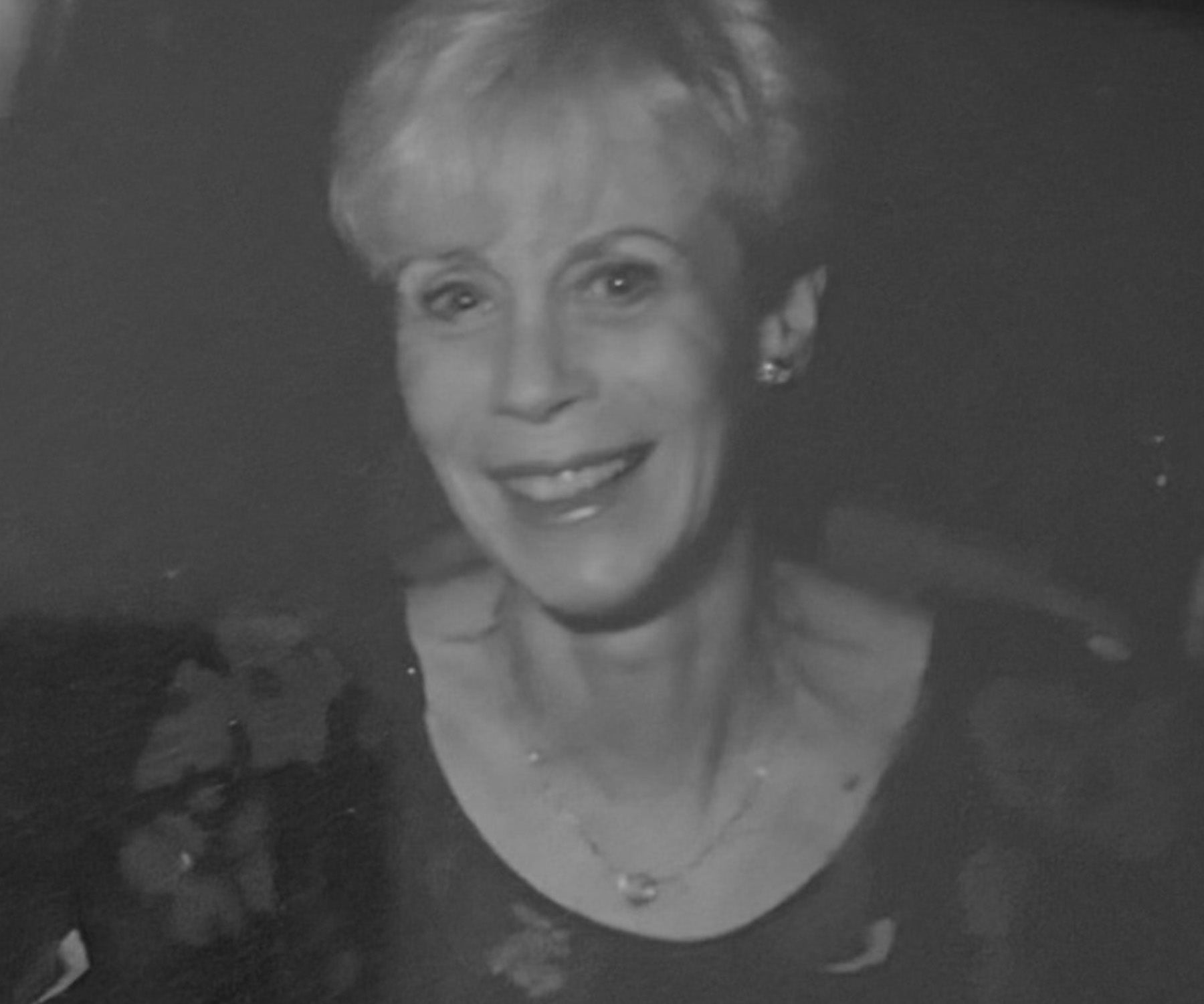 Black and white portrait of a smiling woman with short hair, wearing a dark top and necklace, against a dark background.