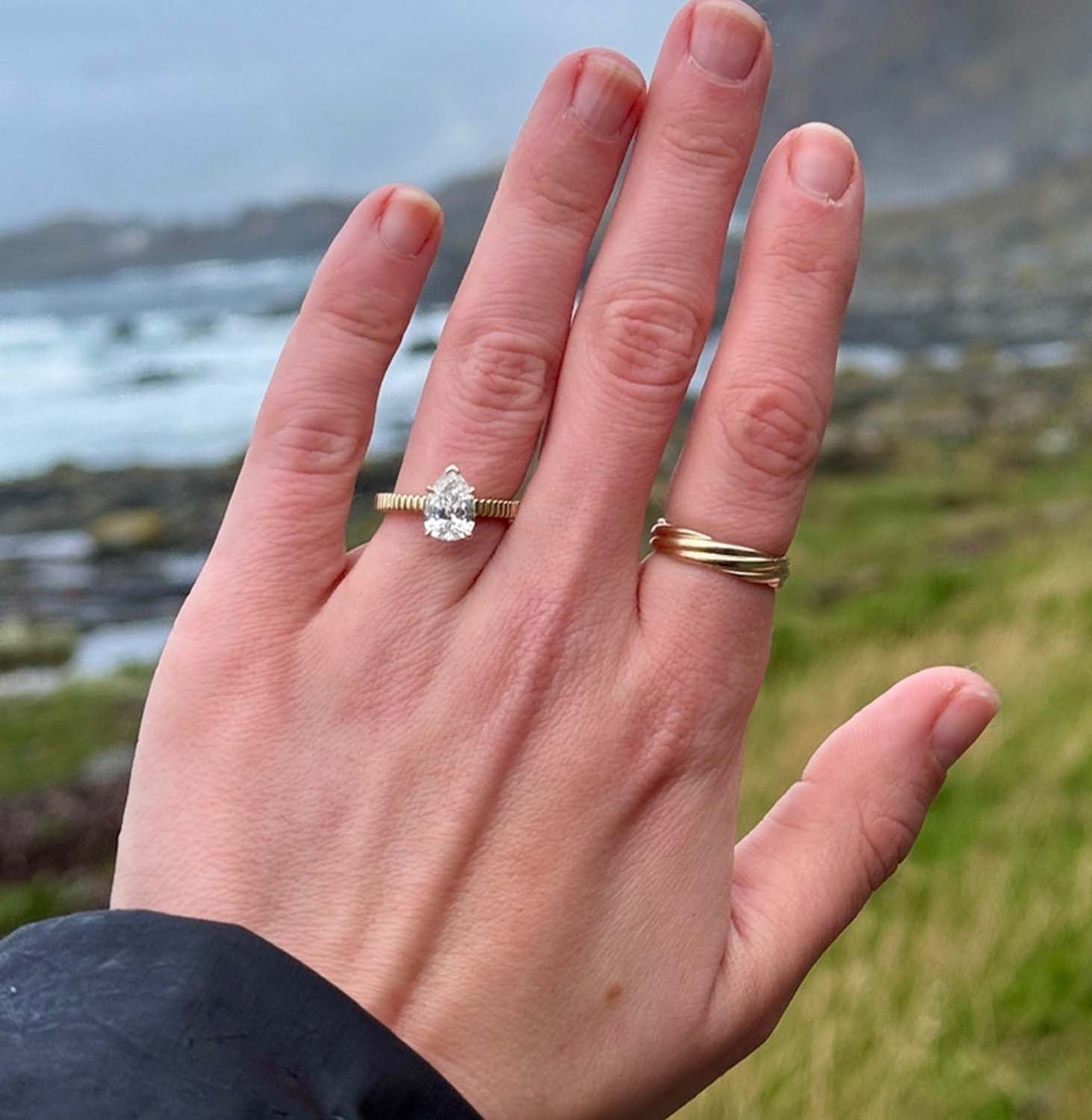 Bethany's hand wearing a Kinn custom diamond engagement ring using an heirloom pear shaped diamond against a scenic background