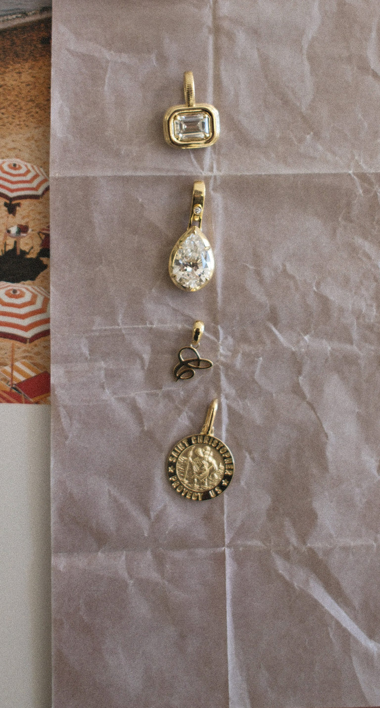 4 Kinn 14k Gold and Diamond Pendants lined up in a row on a light brown crumpled paper background and a beach photo