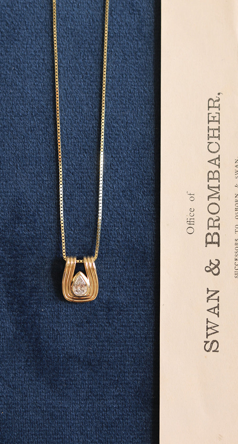 A gold Kinn Unbridled Alyssa pendant necklace with a teardrop-shaped clear gemstone and curved gold settings, displayed on a textured navy blue fabric next to a cream-colored card printed with 'SWAN & BROMBACHER'.