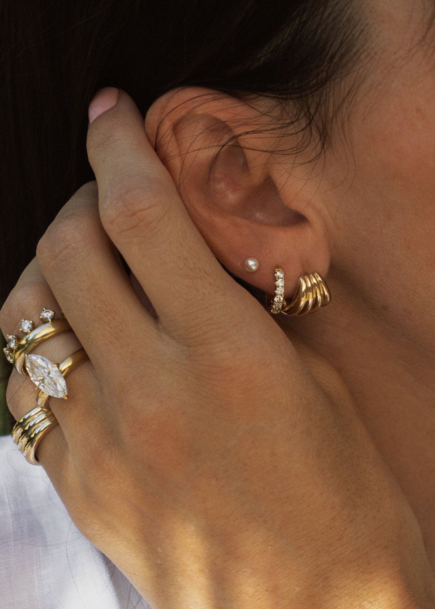 close up of a woman's ear wearing a Kinn 14k Gold Pearl Stud Earring layered with a Gaia Scallop Earring and wearing gold and diamond rings on her hand 