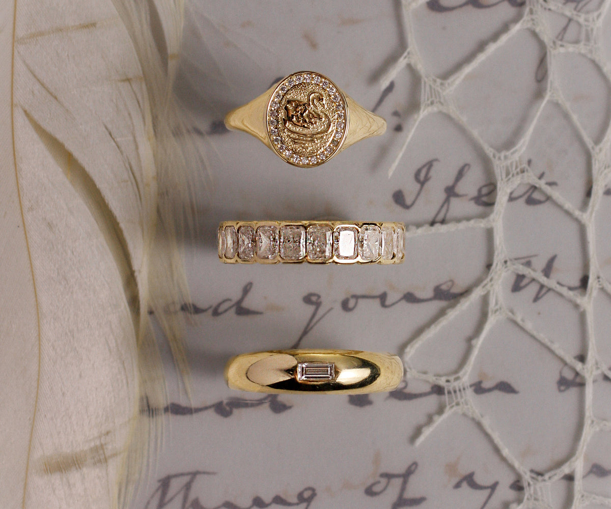 Three gold rings displayed vertically on a vintage-style background with cursive script, lace, and a feather. The top ring is a signet-style ring with a swan motif and clear stones; the middle ring is a band with rectangular clear stones; the bottom ring is a slim gold band with a small clear rectangular stone.