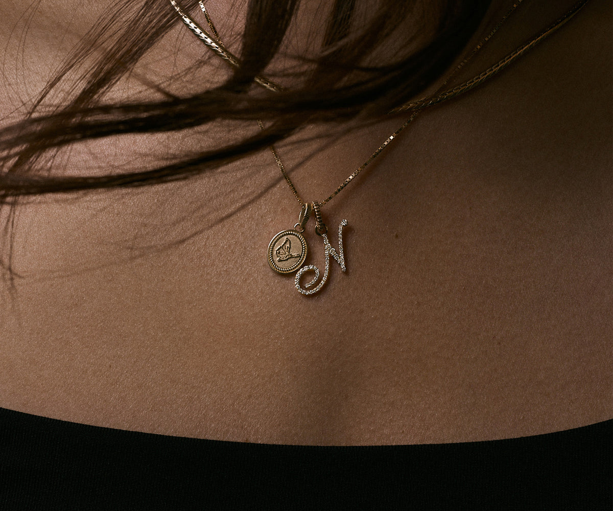 A close-up of a person's chest wearing a delicate gold necklace with a circular initial pendant and a cursive initial 'N' charm, partially covered by dark hair.