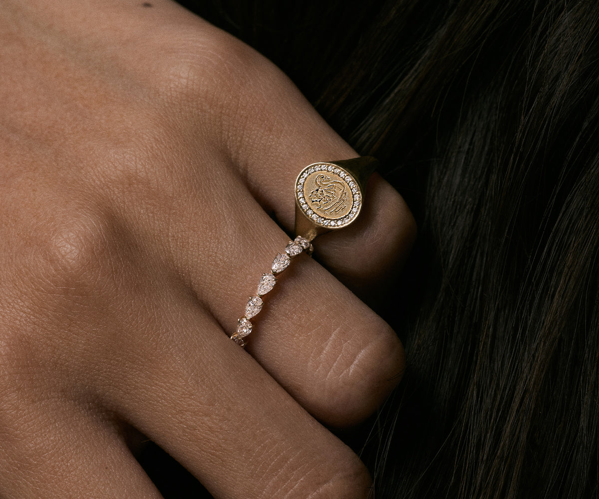 A close-up of a hand wearing two rings: a gold signet-style ring with a circular engraved face and a delicate rose-gold band set with small clear stones, against dark hair.