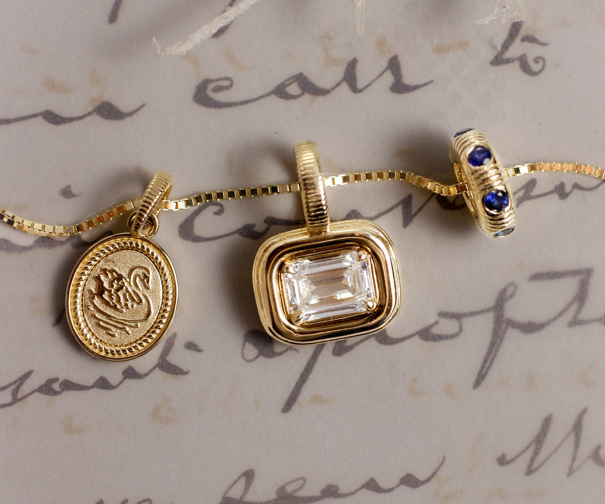 A gold necklace with three charms displayed on a beige background with cursive handwriting. The charms include a round engraved medallion, a rectangular clear gemstone pendant, and a small bead with blue stones.