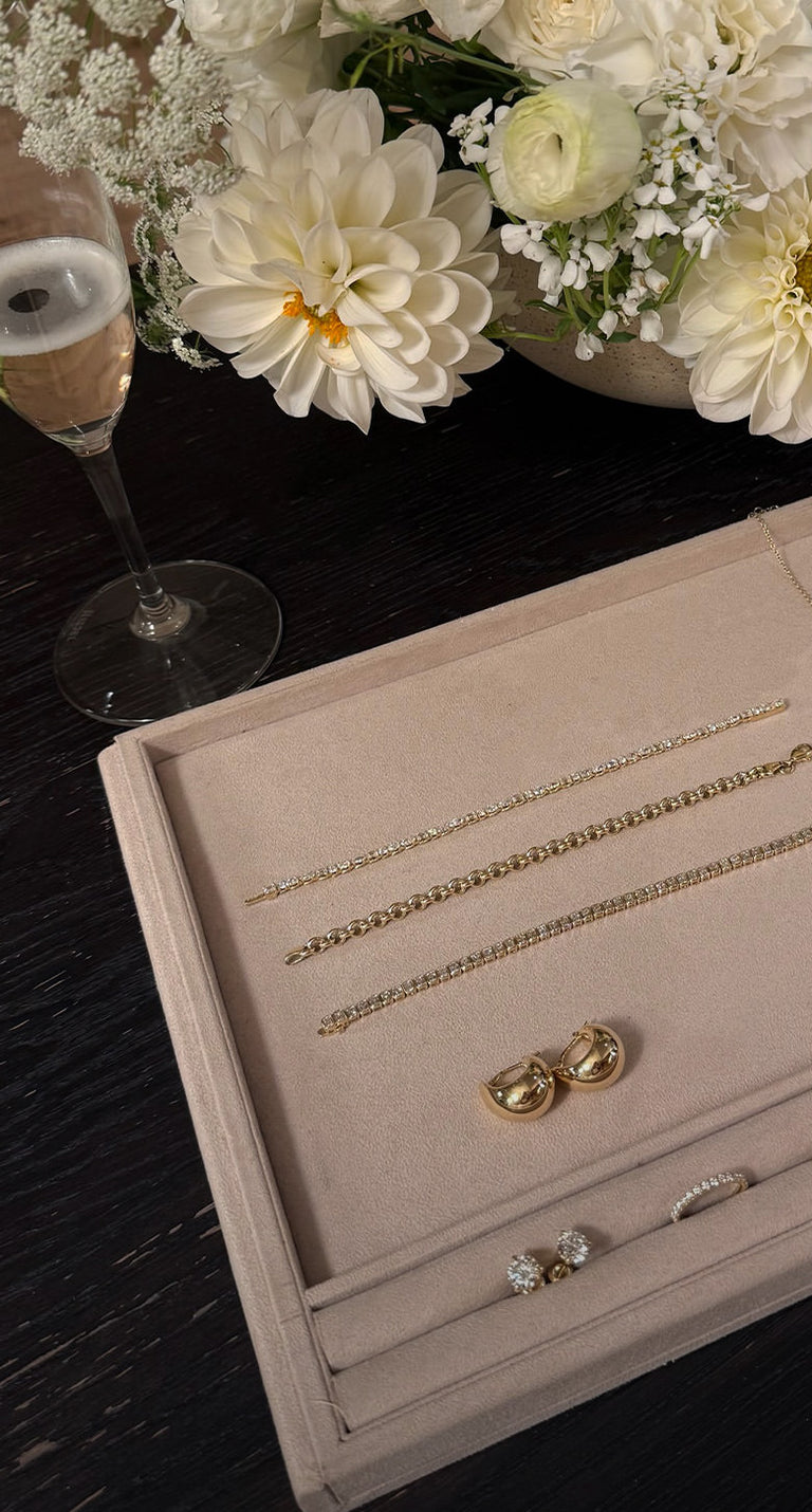 Kinn 14k gold and diamond jewelry in a beige box accompanied by a glass of champagne and flowers on a dark surface.