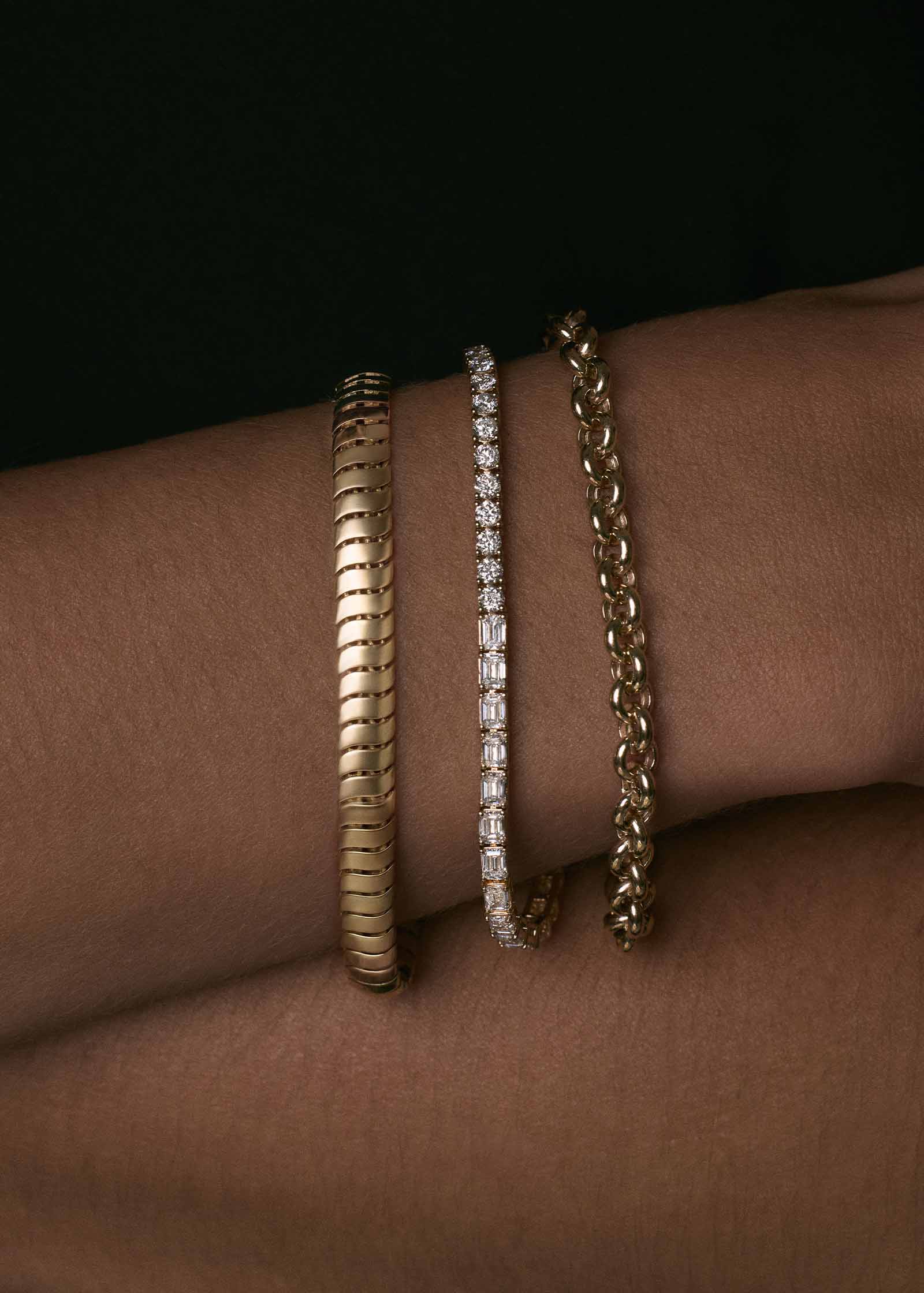close up of a woman's wrist wearing a Kinn x Prince 14k Gold Billie Half Round Emerald Tennis Bracelet stacked with a Cobra Chain and Matis Rolo Chain bracelet