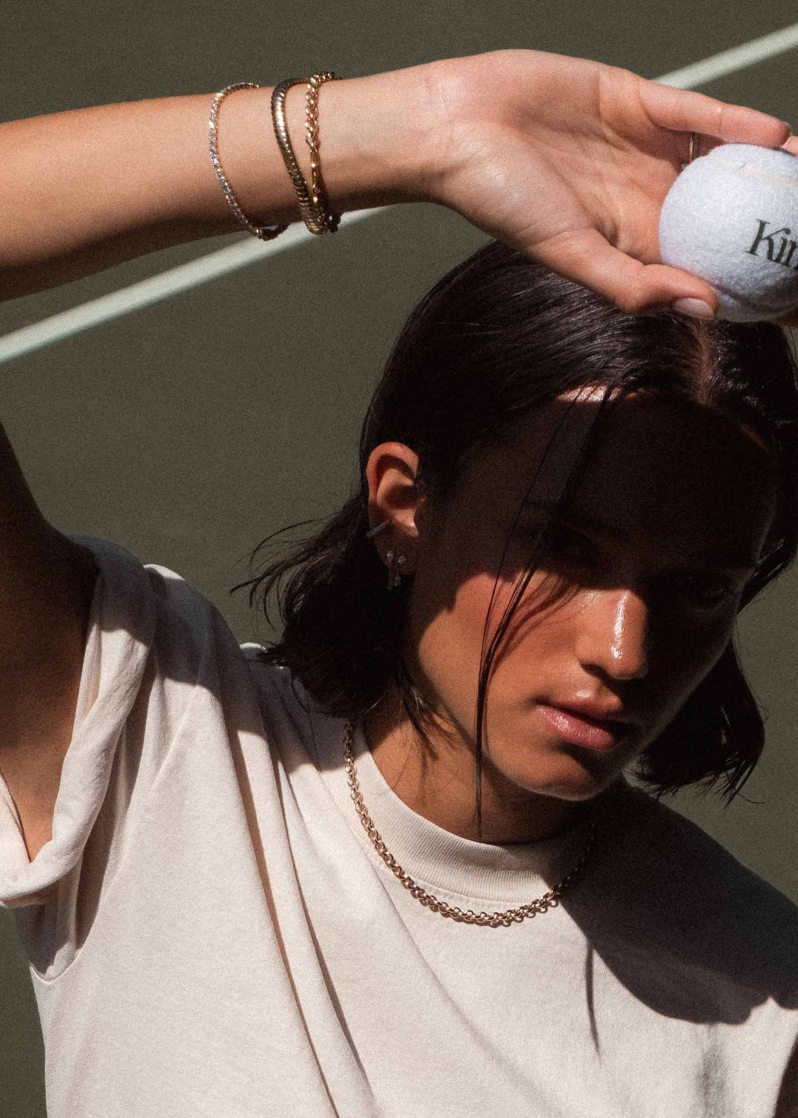 woman with her hand raised holding a tennis ball wearing a Kinn x Prince 14k Gold Billie Half Round Emerald Tennis Bracelet stacked with a Cobra Chain and Matis Rolo Chain bracelet