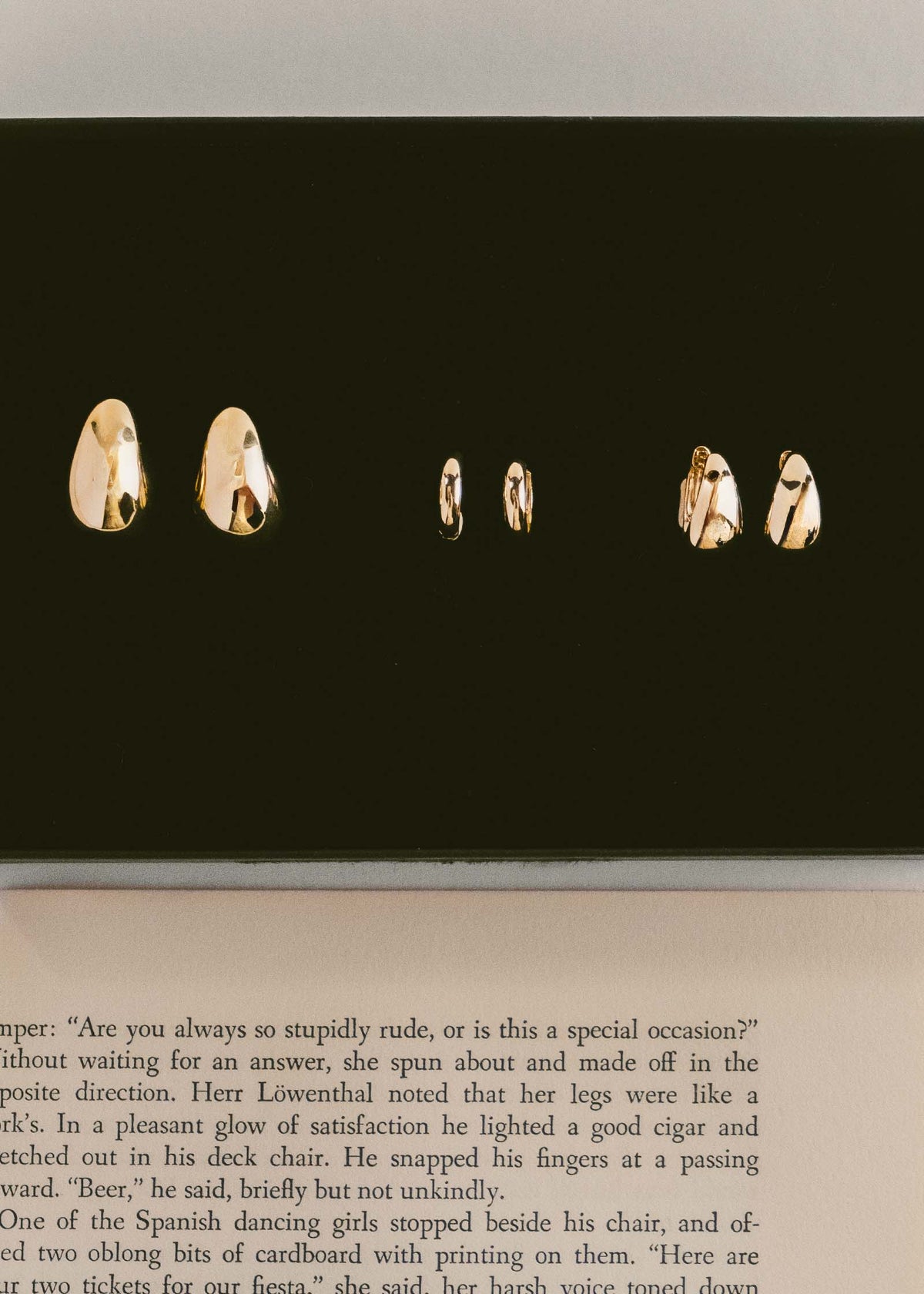 A Kinn 14k Gold Bold Teardrop Hoop Earrings laid flat with a pair of Classic Huggie Earrings and a pair of Petite Bold Teardrop Hoop Earrings in a row against a  velvet background next to a book