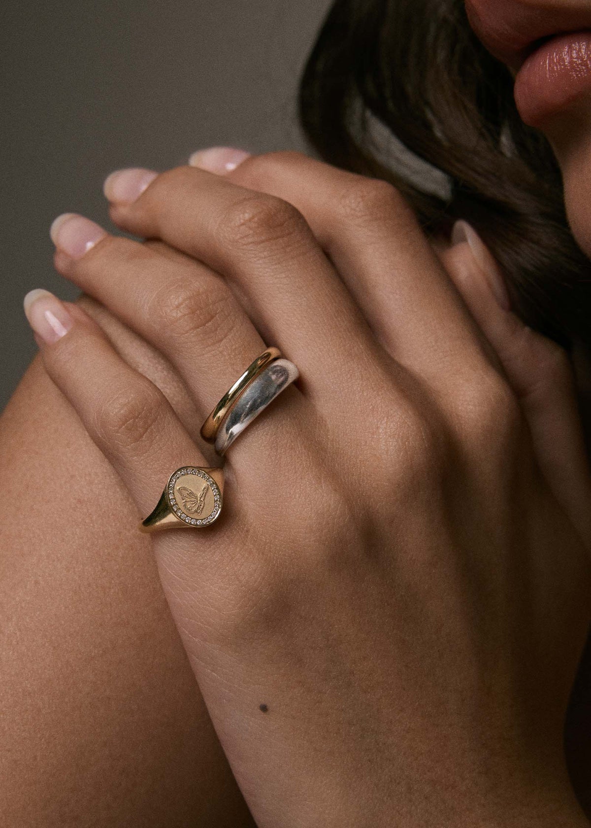 A close-up of a person's hand resting on her shoulder wearing a Kinn 14k Gold Butterfly Dream Diamond Signet Ring on her pinky and a Dare to Love Dome Ring stack on her ring finger