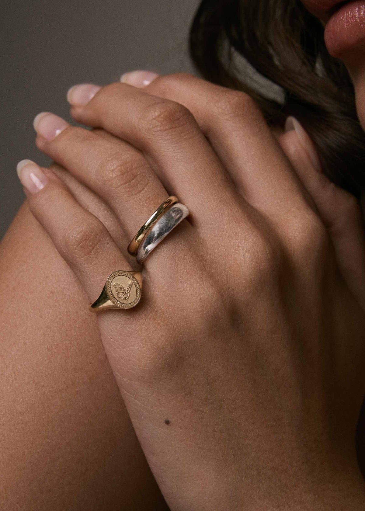 A close-up of a person's hand resting on her shoulder wearing a Kinn 14k Gold Butterfly Dream Signet Ring on her pinky and a Dare to Love Dome Ring stack on her ring finger