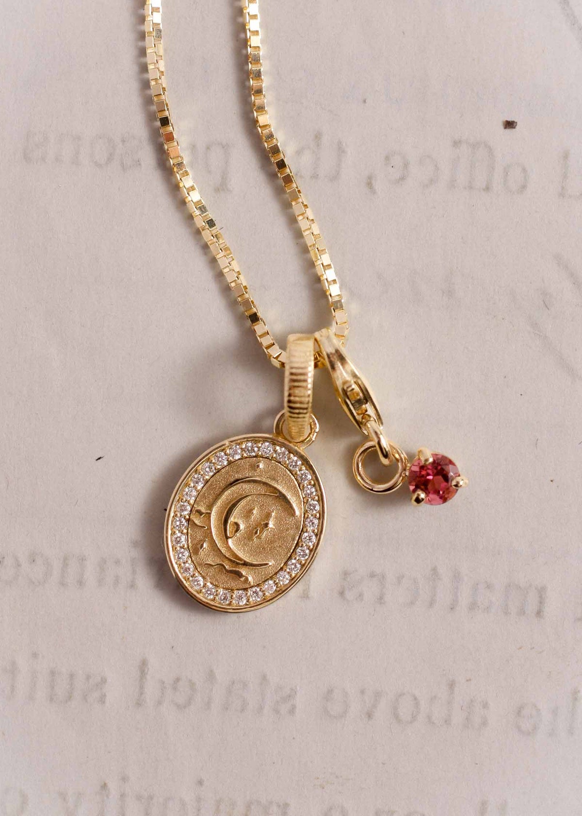 A close up of a Kinn 14k Gold Crescent Dream Diamond Pendant and a Ruby Maeum Charm hanging off a box chain and laying flat against a piece of paper