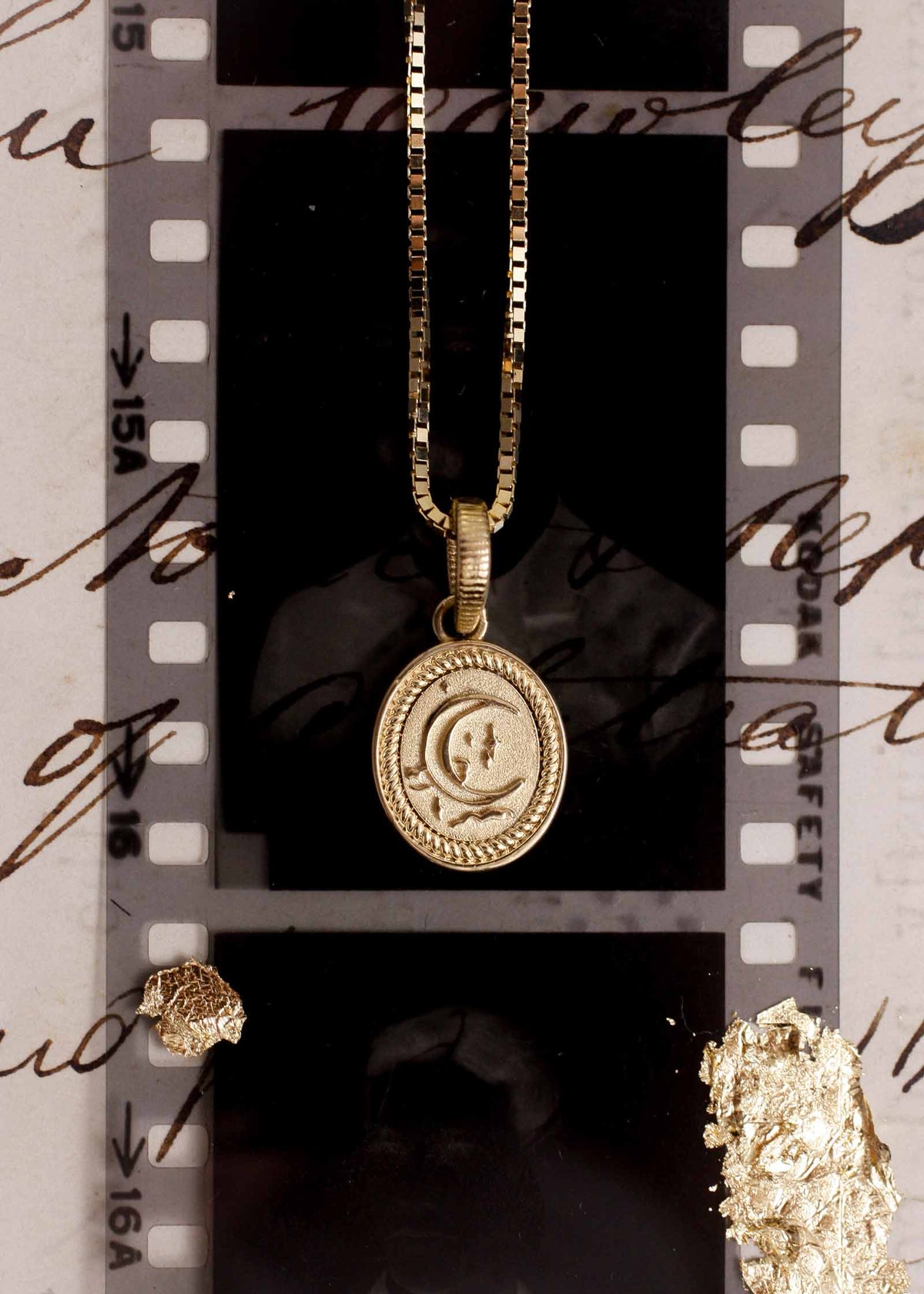 A Kinn 14k Gold Crescent Dream Pendant on a box chain necklace laying flat against a strip of film
