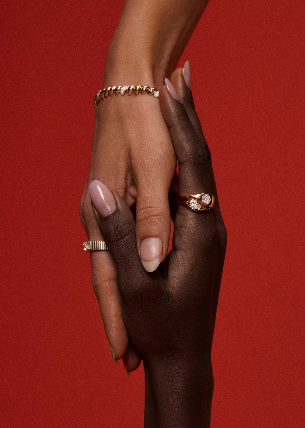 Two hands clasped together against a solid red background, each wearing Kinn gold and diamond jewelry. The hand on the left has a Pave Diamond Petite Hampshire House bracelet and a Solis Ribbed Ring Oval Diamond, while the hand on the right wears a Kinn 14k Gold Gigi Toi et Moi Diamond Ring