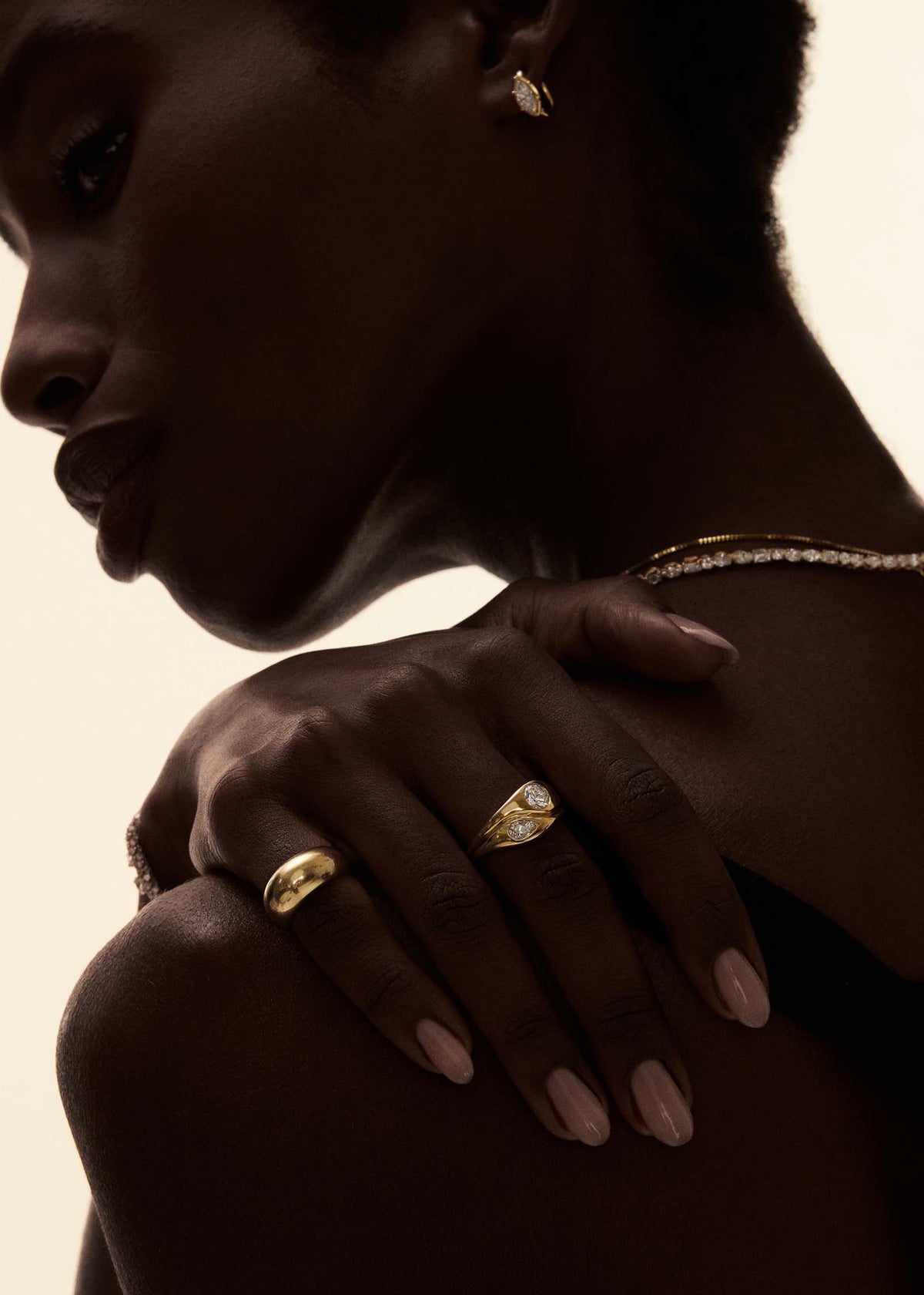 A close-up lifestyle shot of a model wearing a Kinn 14k Gold Gigi Toi et Moi Diamond Round Pear Ring and a Dare to Love Bold Dome Ring on her pinky.  The image is shot in dramatic, warm, low-key lighting against a solid cream background.