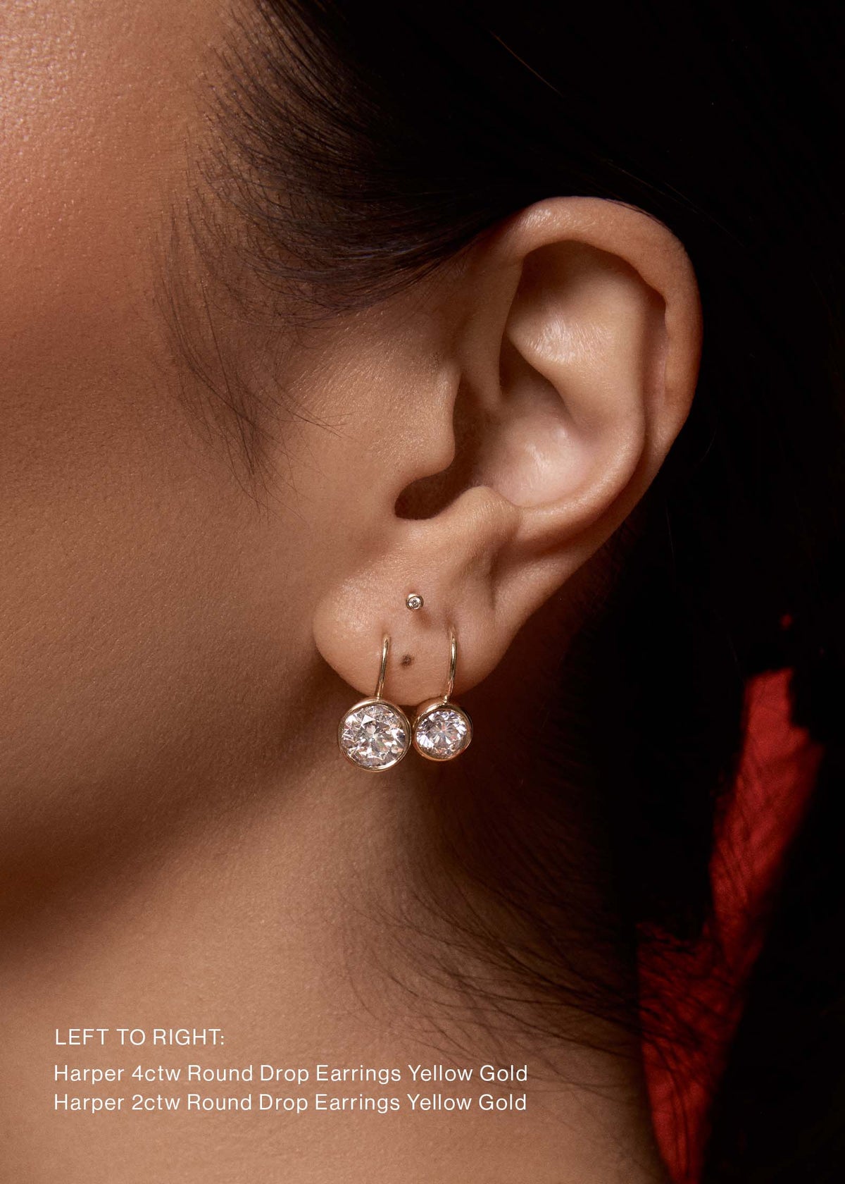 A woman wearing Kinn 14k Yellow Gold Harper 4ctw Round Drop Earrings layered with Harper 2ctw Round Drop Earrings in her second piercing against a red background and with text at the bottom left of the image listing the product names