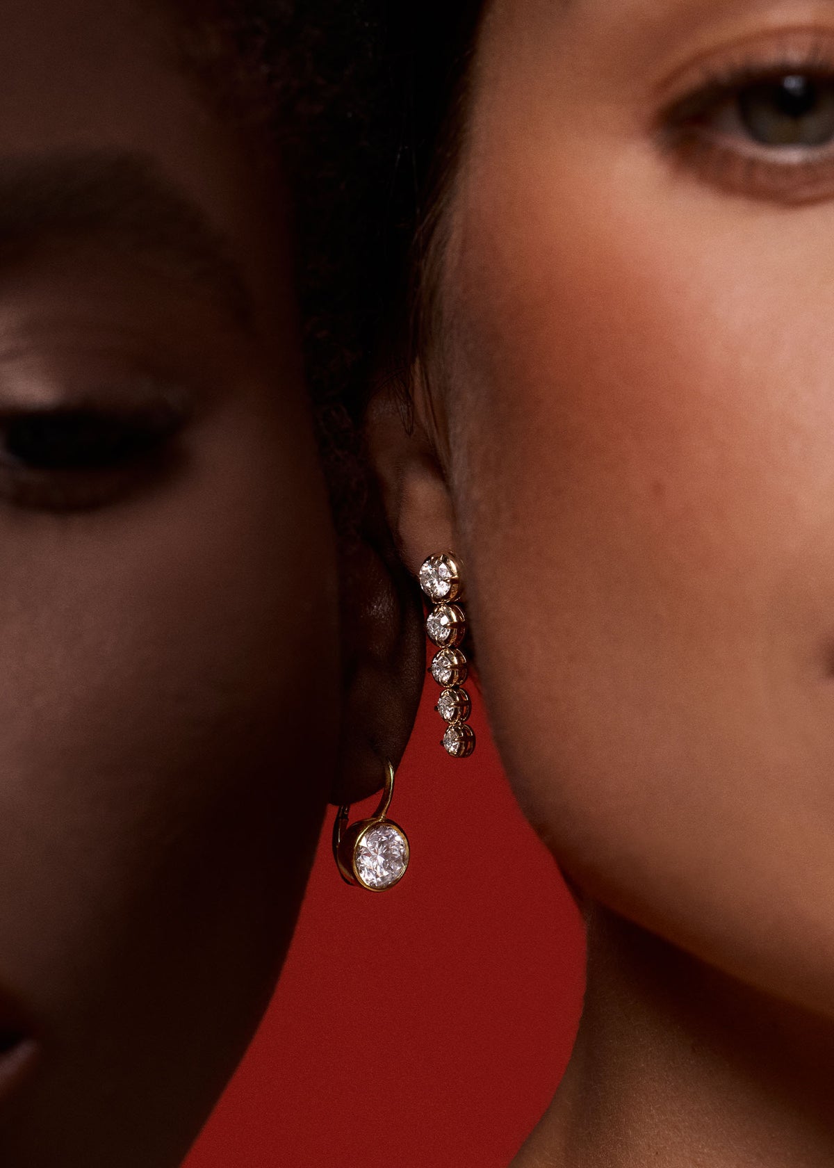 Two woman with their faces next to each other against a red background with one woman wearing a Kinn 14k Yellow Gold Harper 4ctw Round Drop Earring and the other wearing a Naomi Graduated Diamond Drop earring