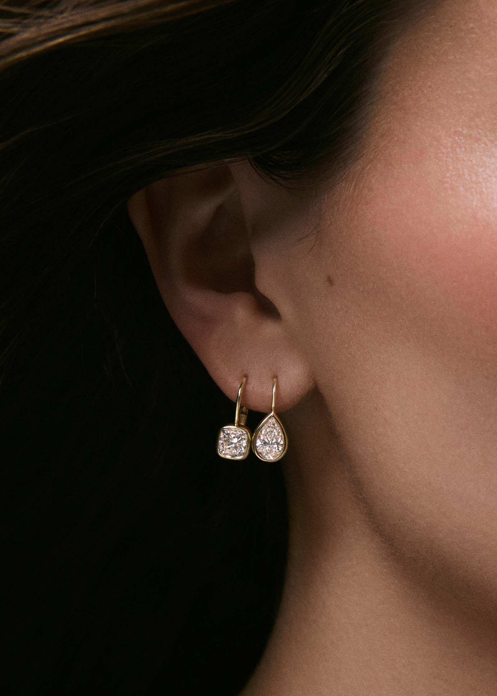 A close up of a woman's ear wearing a Kinn 14k Gold Harper 2ctw Cushion Cut Diamond Drop Earring in her second piercing and a Harper 2ctw Pear Cut Diamond Drop Earring in her first