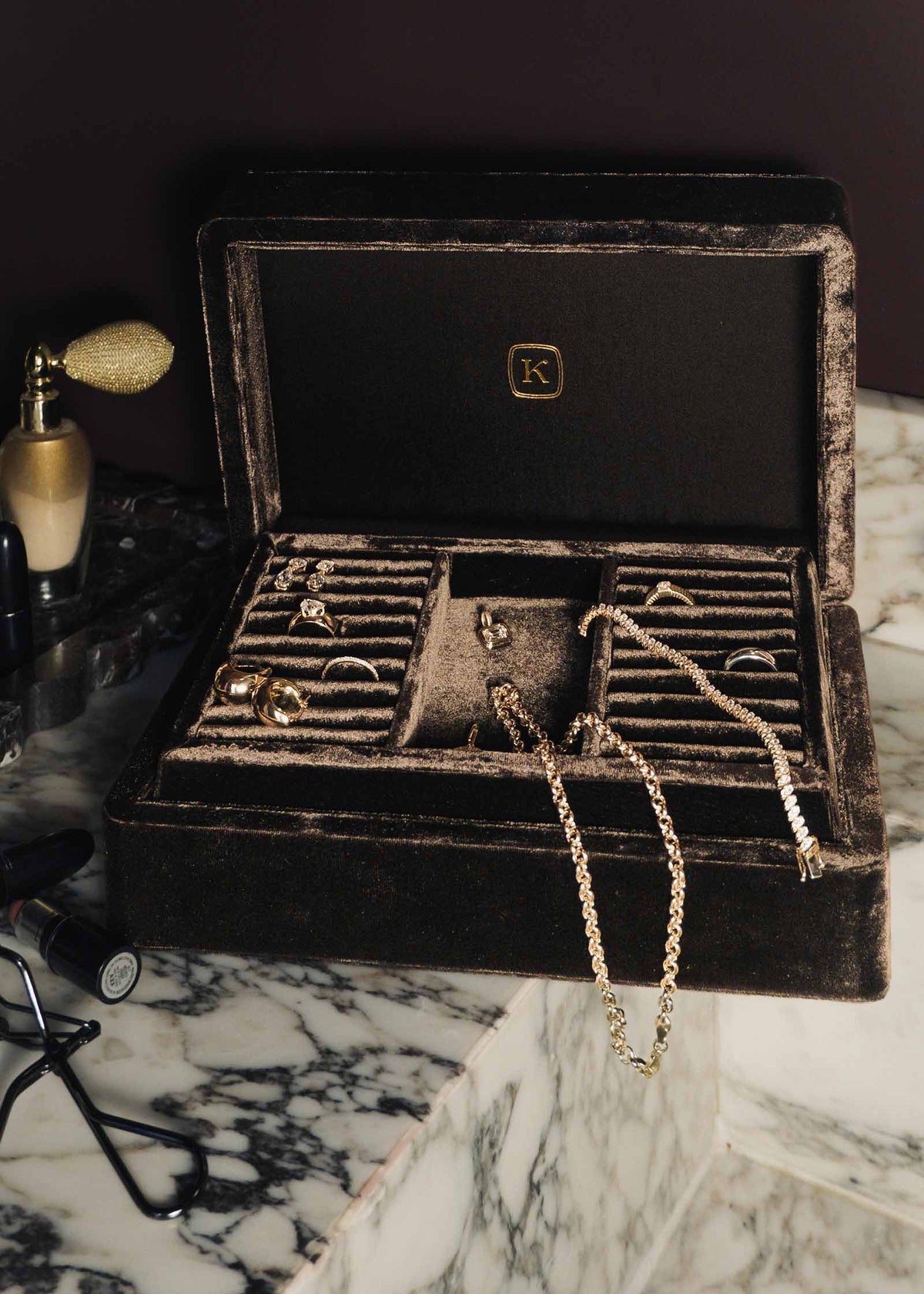 An open The Keepsake Jewelry Box by Kinn Studio with multiple compartments holding gold-toned rings and chains, placed on a marble surface against a dark background.