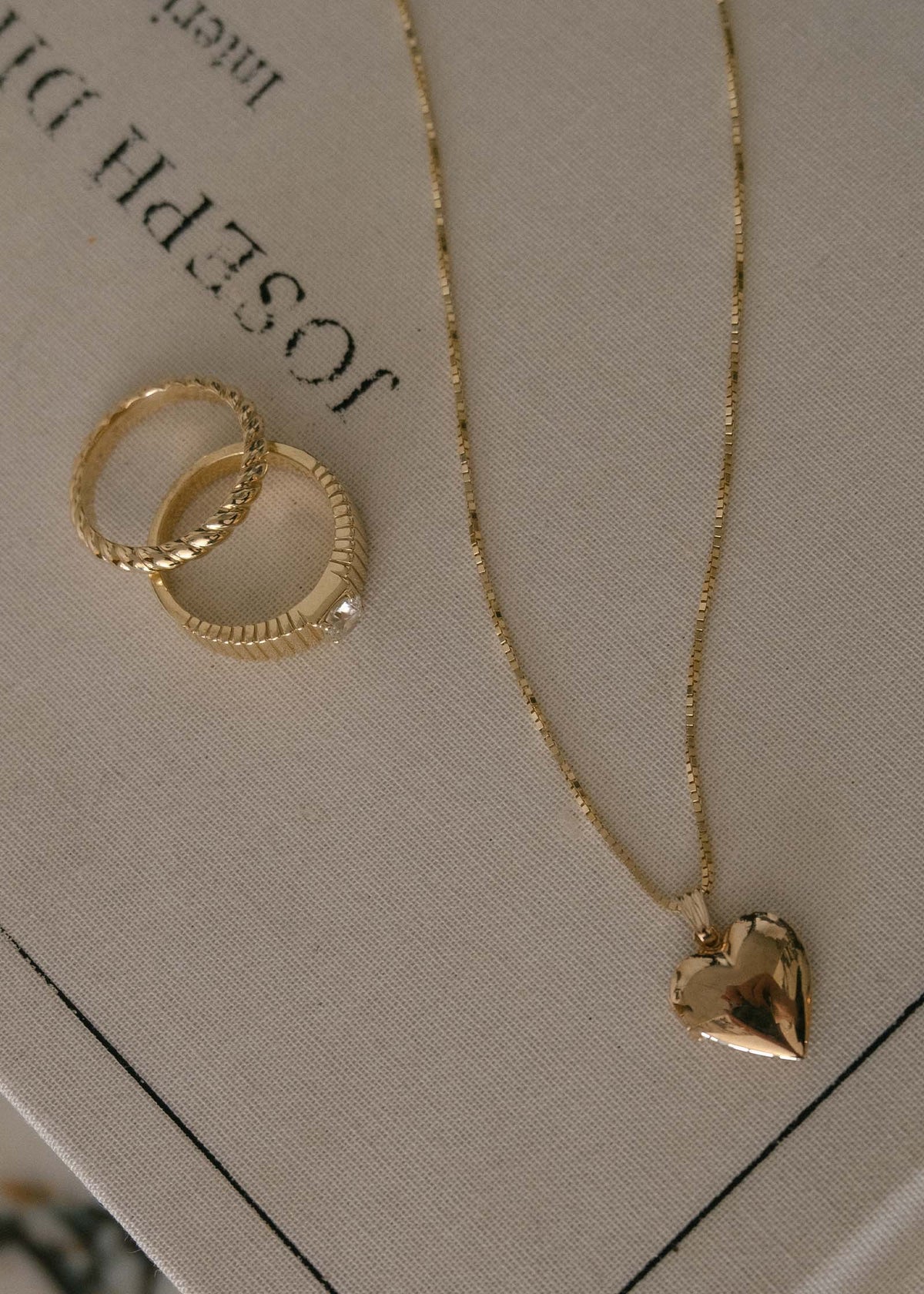 A Kinn 14k Gold Maison Heart Locket Pendant on a box chain laying flat on a book next to a Solis Ribbed Round Diamond .5 ct Ring and Claudine Twist Ring