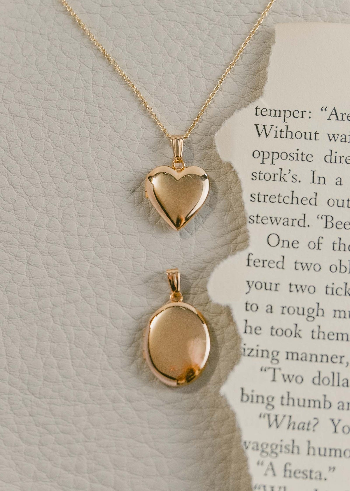 A Kinn 14k Gold Maison Heart Locket Necklace laying flat with a Maison Oval Locket next to a torn up book page against off white leather