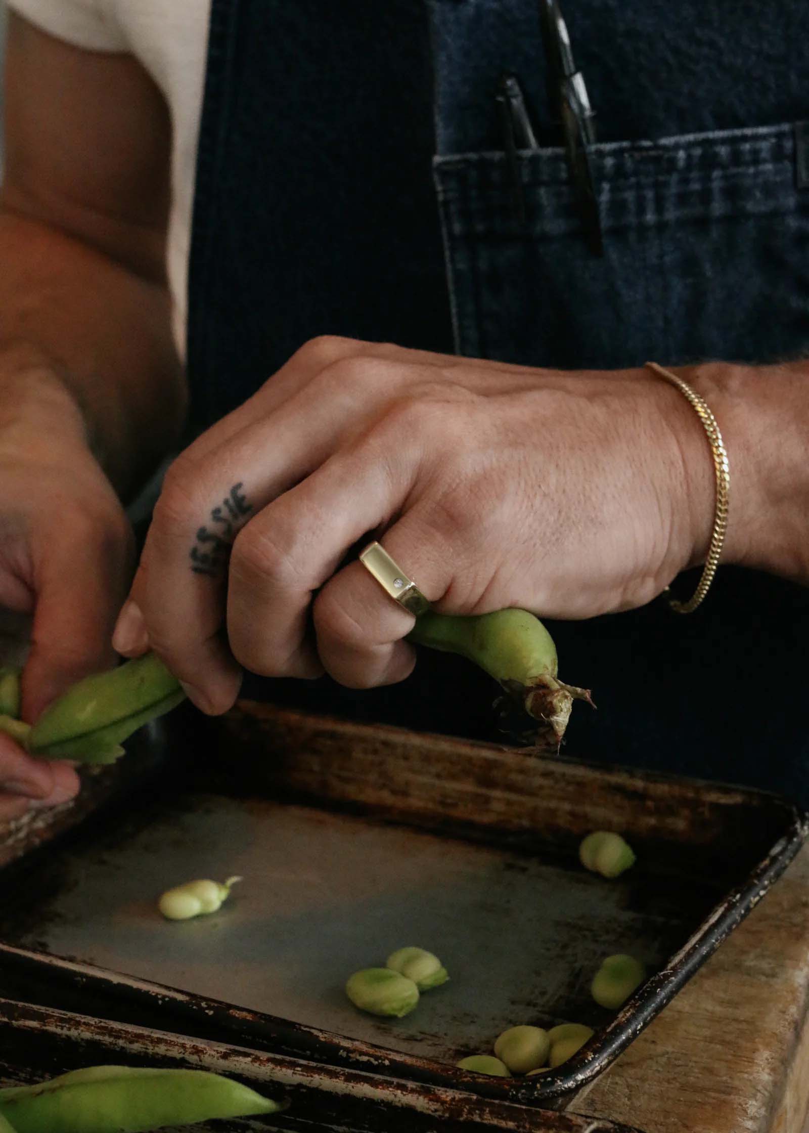 Close up of a man peeling lima beans while wearing a Kinn Studio 14k Gold Benedict Men’s Signet Diamond Ring and a Petite Familia Cuban Chain Bracelet
