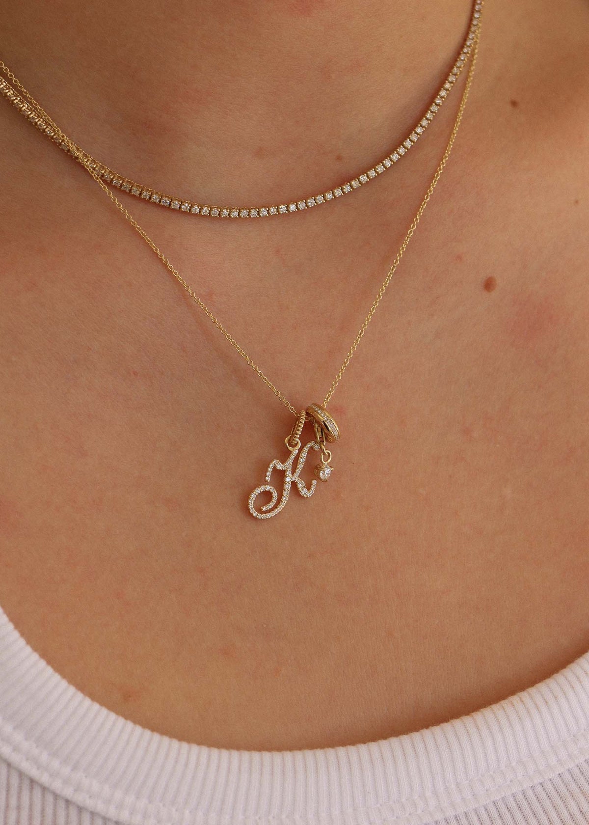 A woman wearing a Kinn 14k Gold Diana Tennis Necklace layered with a Micro rolo chain with a Pavé Diamond Love Letter K Charm and Noelle Diamond Charm hanging from the chain
