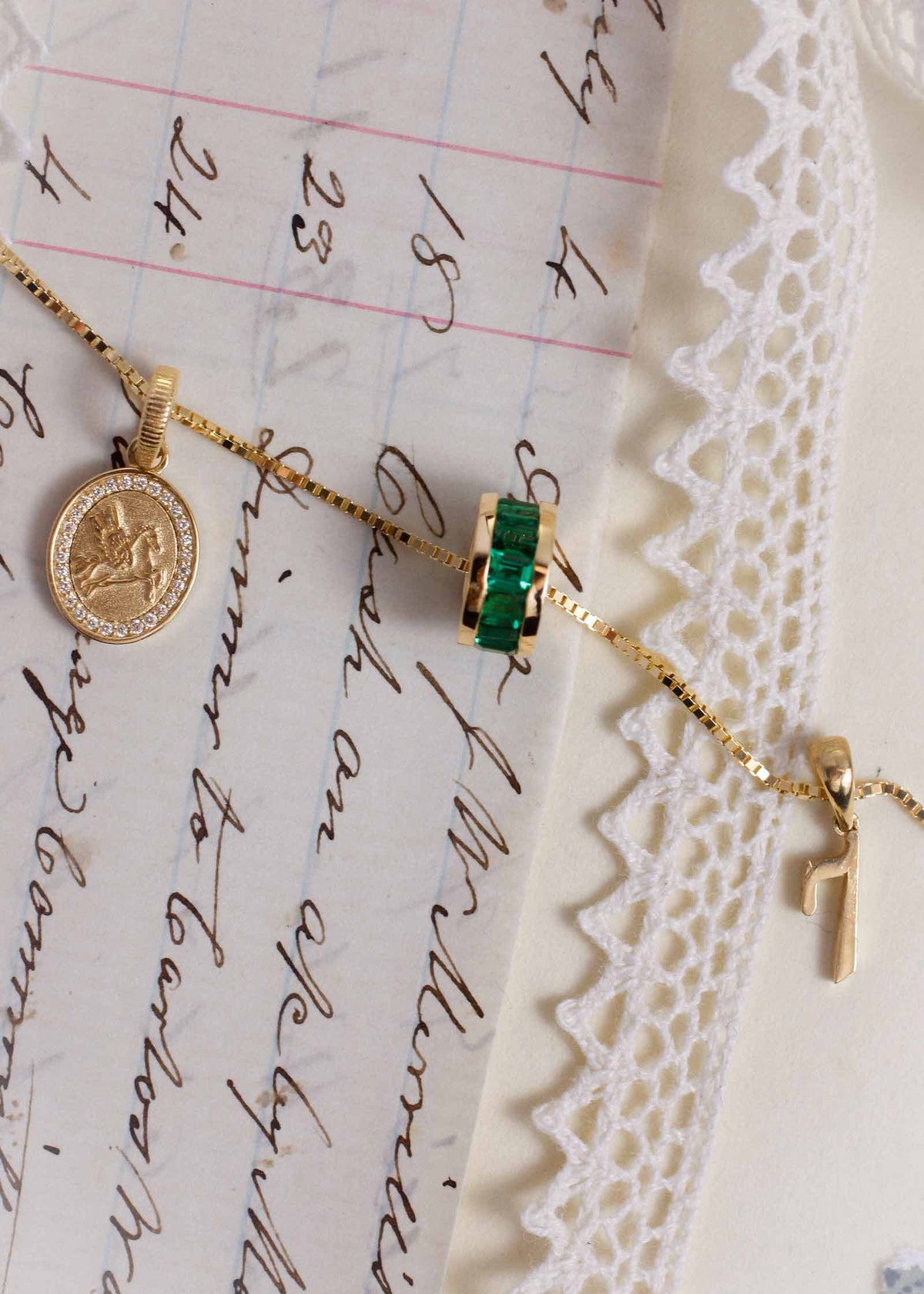 A box chain laying flat with a Kinn 14k Gold Pegasus Dream Diamond Pendant, Milestone Baguette Emerald Charm, and Lucky Number 7 charm hanging from the chain