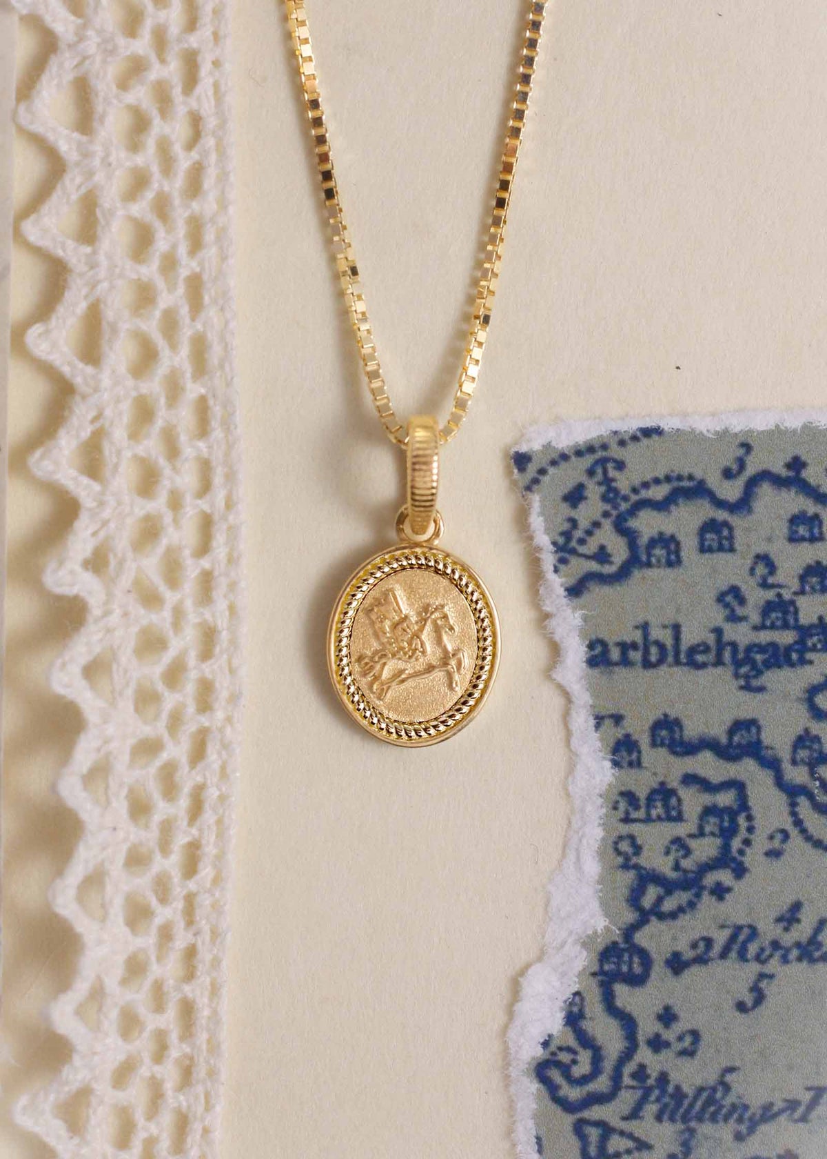 A Kinn 14k Gold Pegasus Dream Pendant on a box chain necklace laying flat against a blue and white background