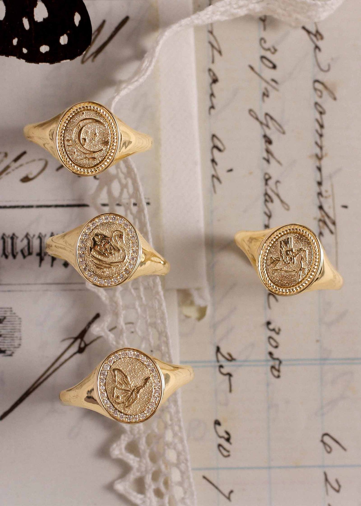 A Kinn 14k Gold Pegasus Dream Signet Ring, Crescent Dream Signet Ring, Swan Dream Diamond Signet Ring, and a Butterfly Dream Diamond Signet Ring sitting upright against a handwritten letter and lace