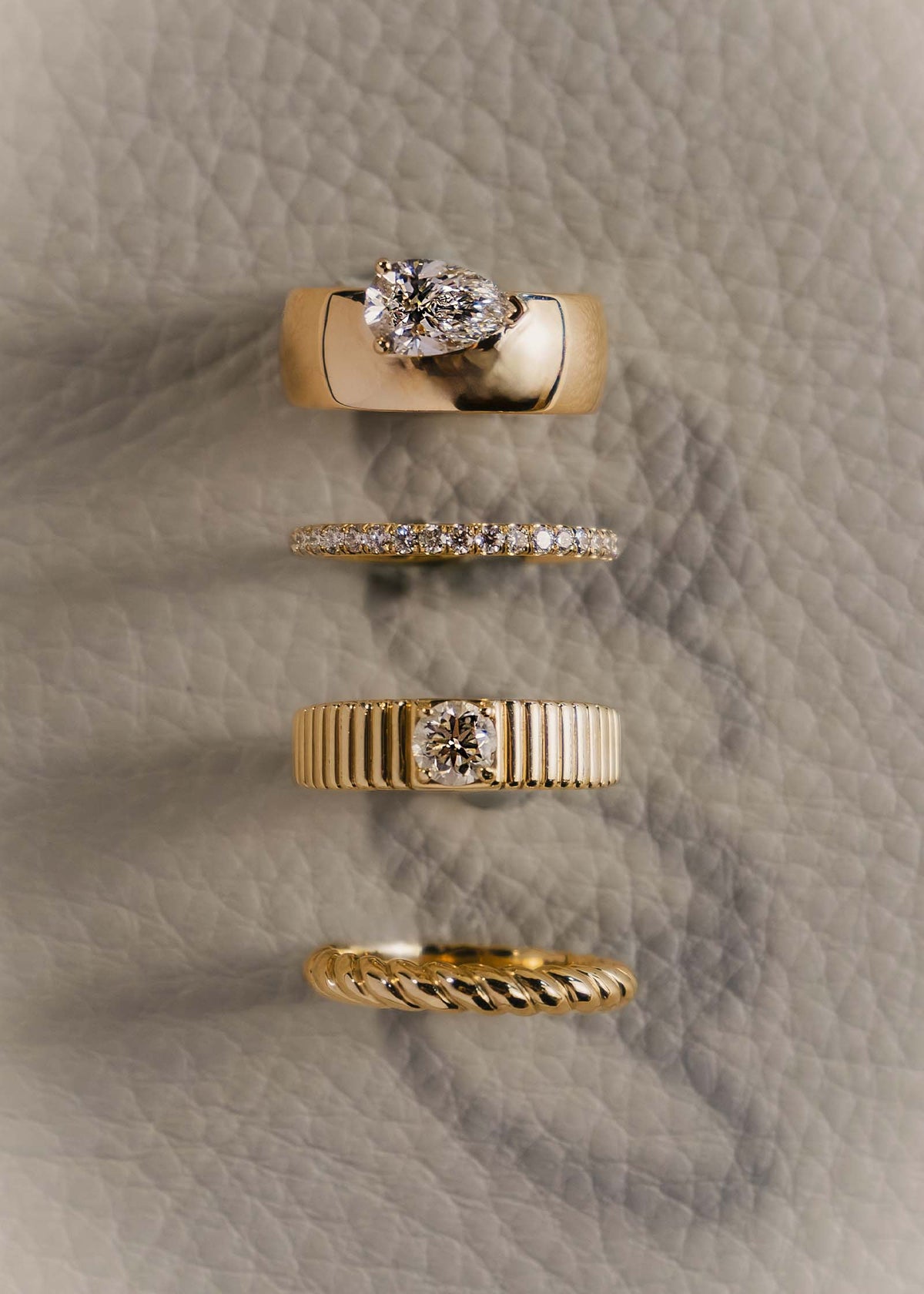 A Kinn 14k Gold Claudine Twist Band Ring, Solis Ribbed Ring Round Diamond 0.5 Carat, Dahlia Pavé Diamond Band, and a Porter Cigar Band Pear Diamond Ring sitting upright in a stacked column against beige leather