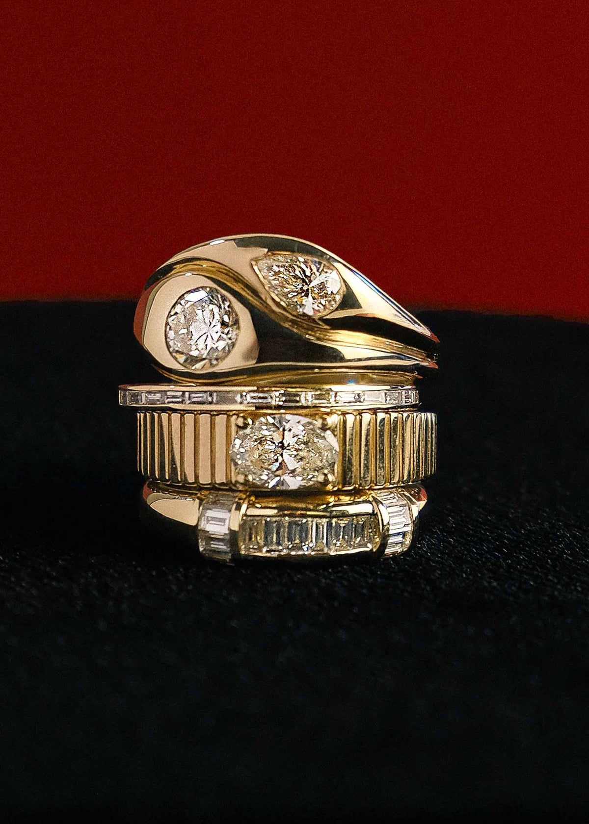 A Kinn Gigi Toi et Moi Ring, 14k Gold Solis Ribbed Ring Oval Diamond, Petite Isabelle Baguette Diamond Ring, and Beatrice Channel Set Baguette Diamond Ring stacked together on black velvet against a red background