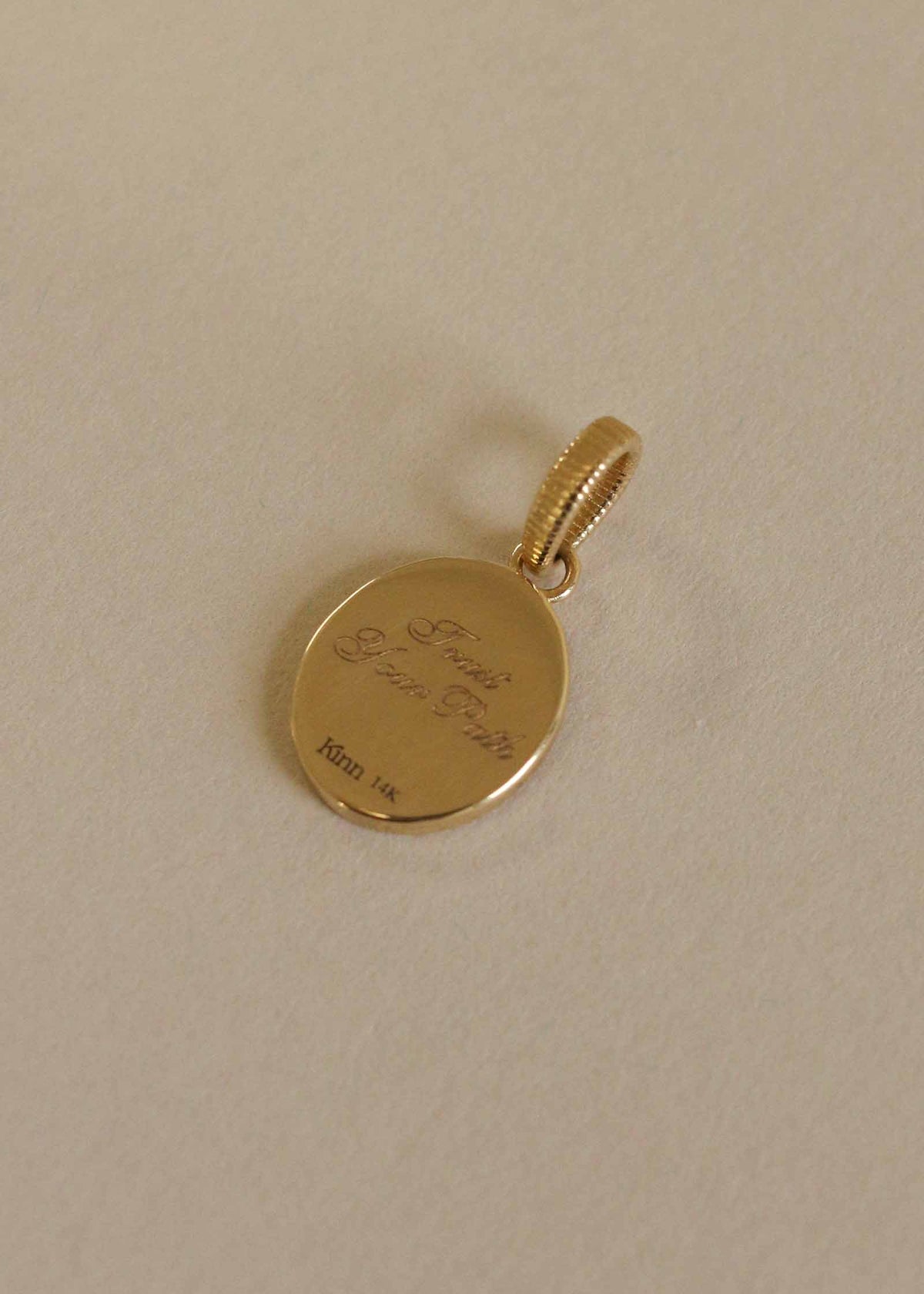 Back of a Kinn 14k Gold Swan Dream Diamond Pendant engraved with "Trust Your Path" in script font laying on a beige background