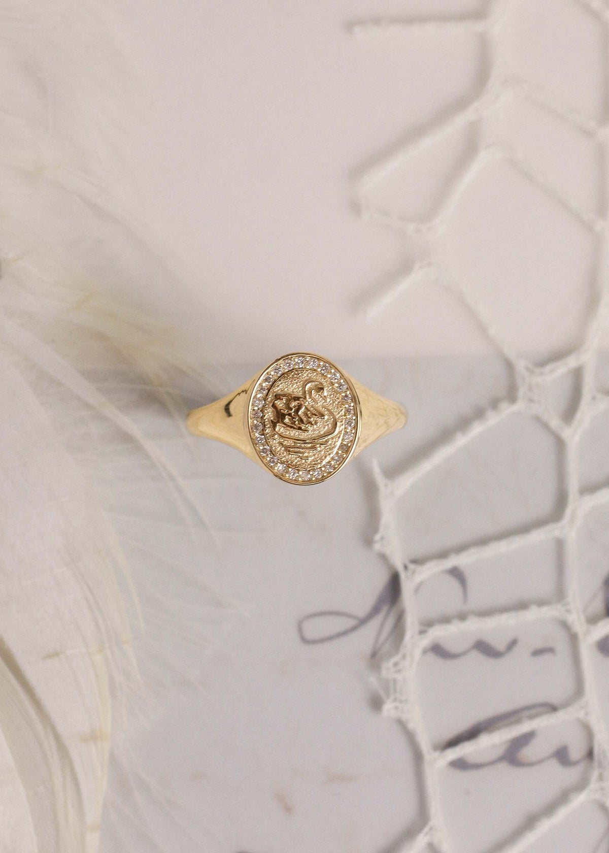 A gold signet ring with a circular medallion featuring a swan motif and small clear stones around the bezel, displayed on a textured white surface with feathers and lace.