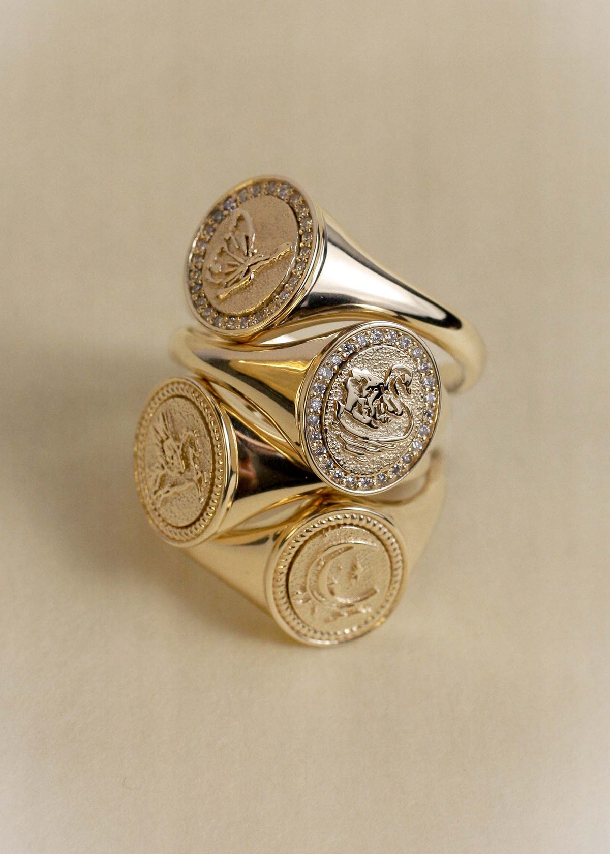 A Kinn 14k Gold Swan Dream Diamond Signet ring stacked with a Butterfly Dream Diamond Signet Ring, Pegasus Dream Signet Ring, and Crescent Dream signet Ring on a warm beige background