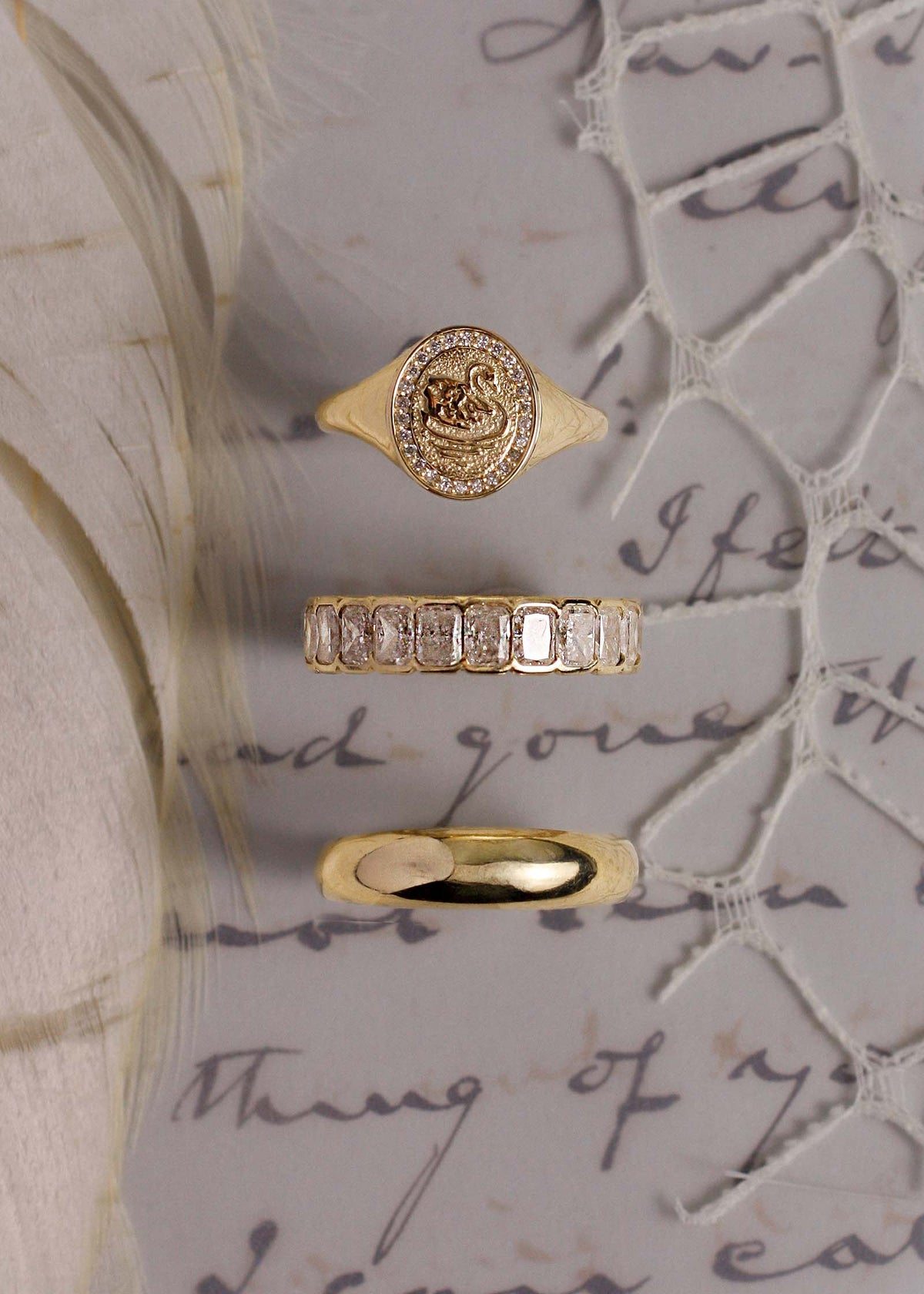 A Kinn 14k Gold Swan Dream Diamond Signet Ring, Jane Radiant Eternity Diamond Ring, and a Dare to love Dome ring siting upright against a handwritten note, feather, and lace