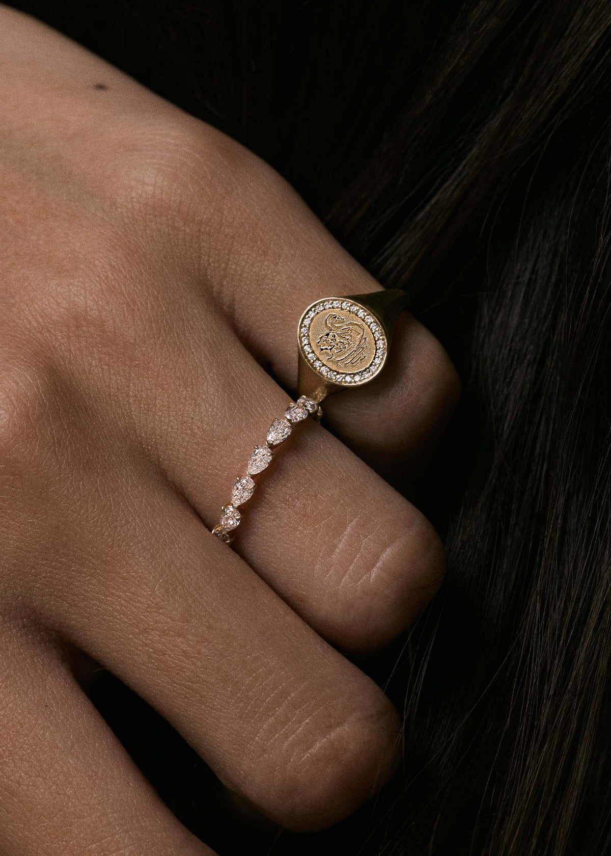 A close up of a woman's hand wearing a Kinn 14k Gold Swan Dream Diamond Signet Ring on her pinky and a Petite Pear Diamond Eternity Ring on her ring finger
