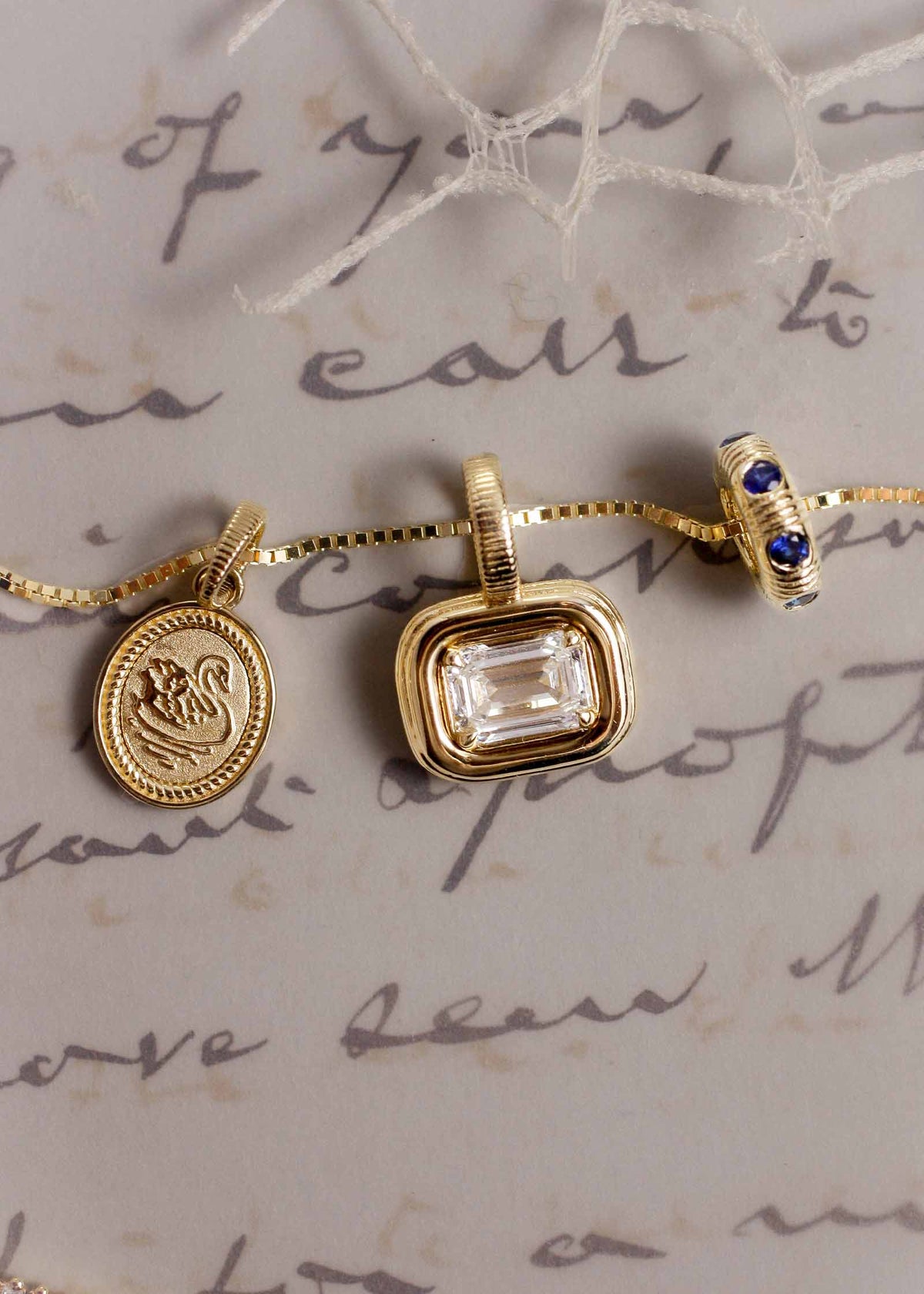 A Kinn Studio 14k Gold Swan Dream Pendant, Margot Diamond Pendant and Blue Sapphire Anniversary Charm on a box chain laying flat against a letter