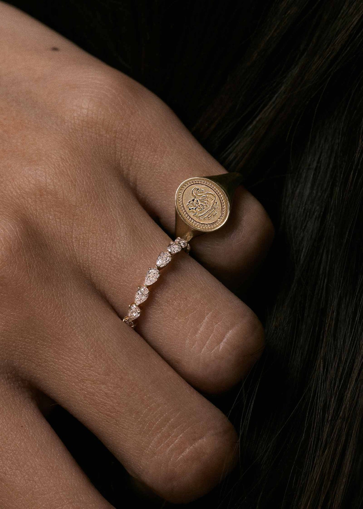 A close-up of a hand wearing a Kinn 14k Gold Swan Dream Signet Ring and Petite Pear Diamond Eternity Ring set against dark hair