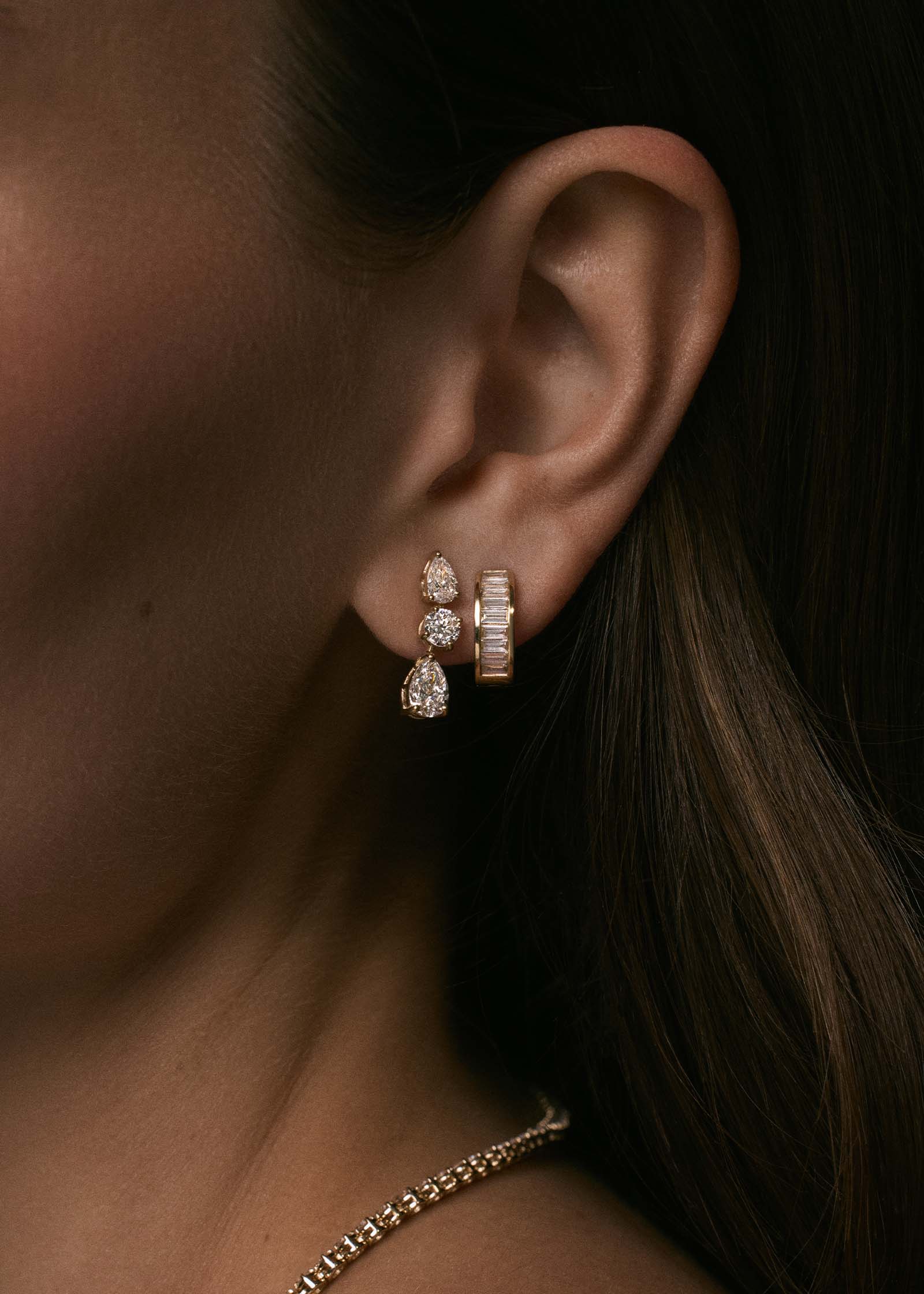Close up of a woman's ear wearing a Kinn 14k Gold Third Time's a Charm Drop Earrings Pear 2.7ctw Diamond layered with a Bobbi Baguette Diamond Huggie Earring in her second piercing