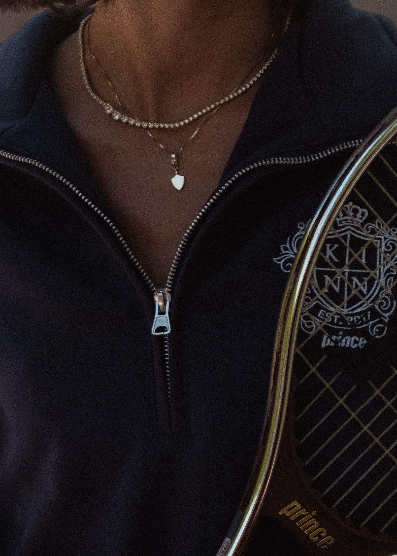 woman in a navy KInn x Prince Quarter Zip Sweatshirt wearing a Kinn 14k Gold Ace Shield Diamond Pendant on a box chain necklace layered with a Naomi Graduated Diamond Tennis Necklace