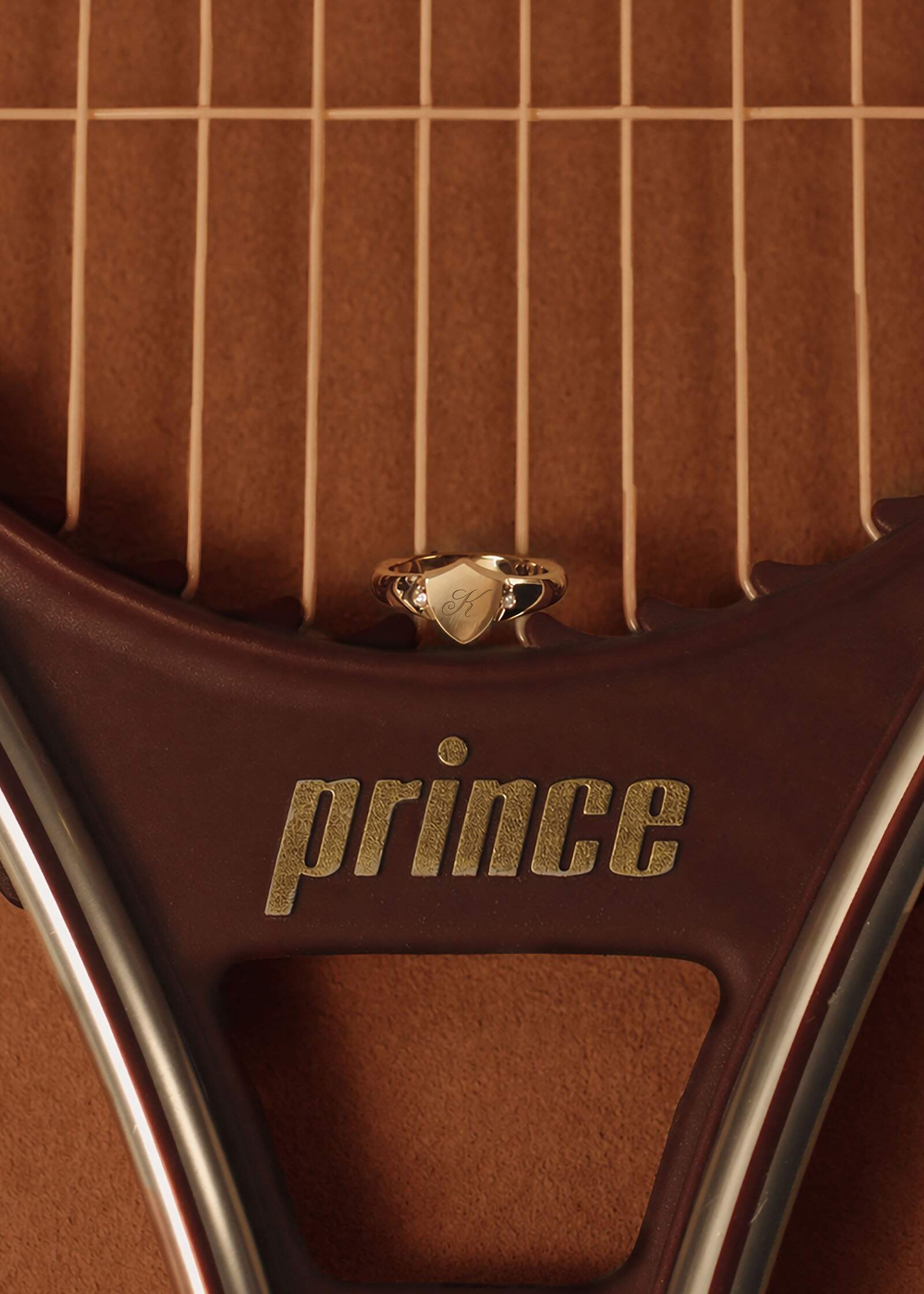 A Kinn x Prince 14k Gold Ace Shield Diamond Signet Ring sitting on top of the strings of a vintage Prince tennis racquet against a clay court background