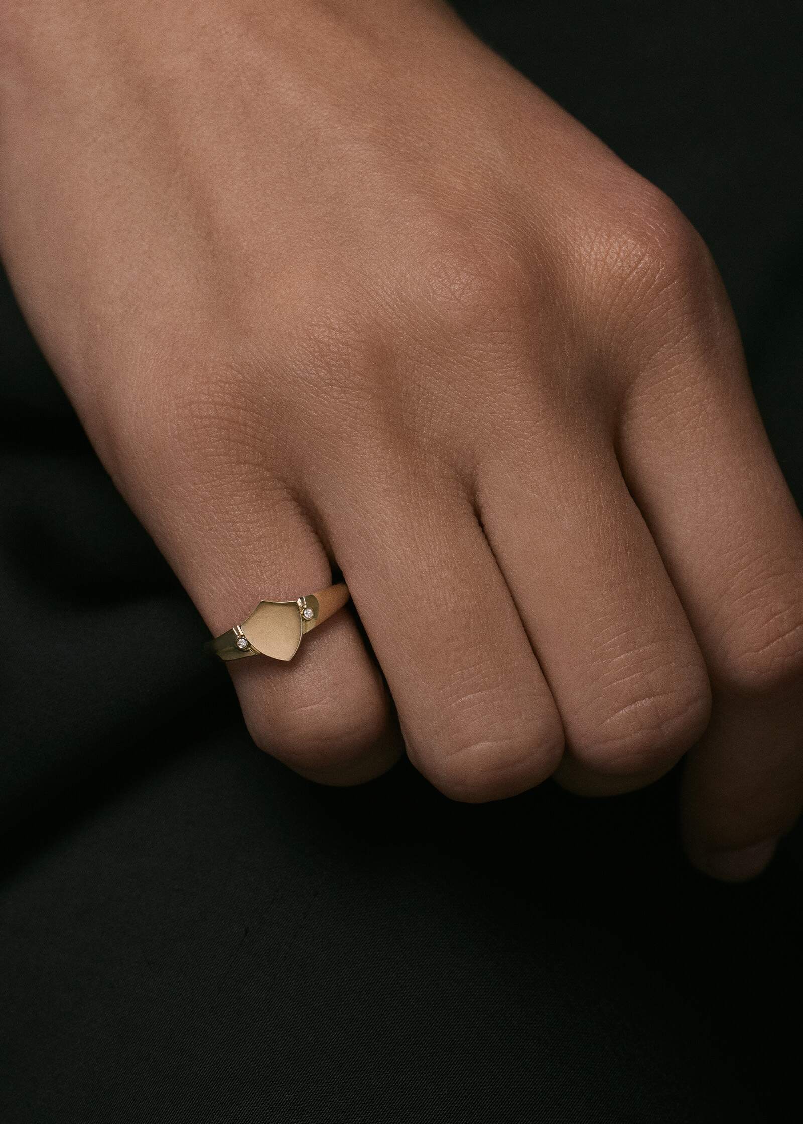 Close up of a hand wearing a Kinn x Prince 14k Gold Ace Shield Diamond Signet Ring on her pinky finger against a black background