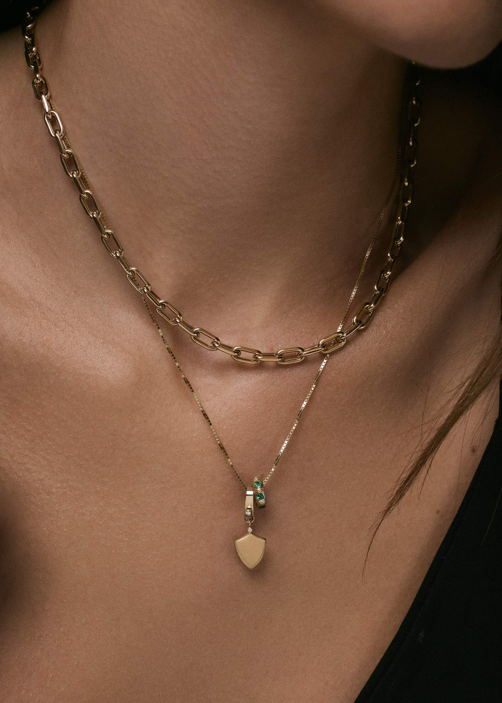 A close up of a woman wearing a Kinn 14k Gold Ace Shield Diamond Pendant and an Anniversary Charm Emerald on a box chain layered with a Mia Link Chain Necklace