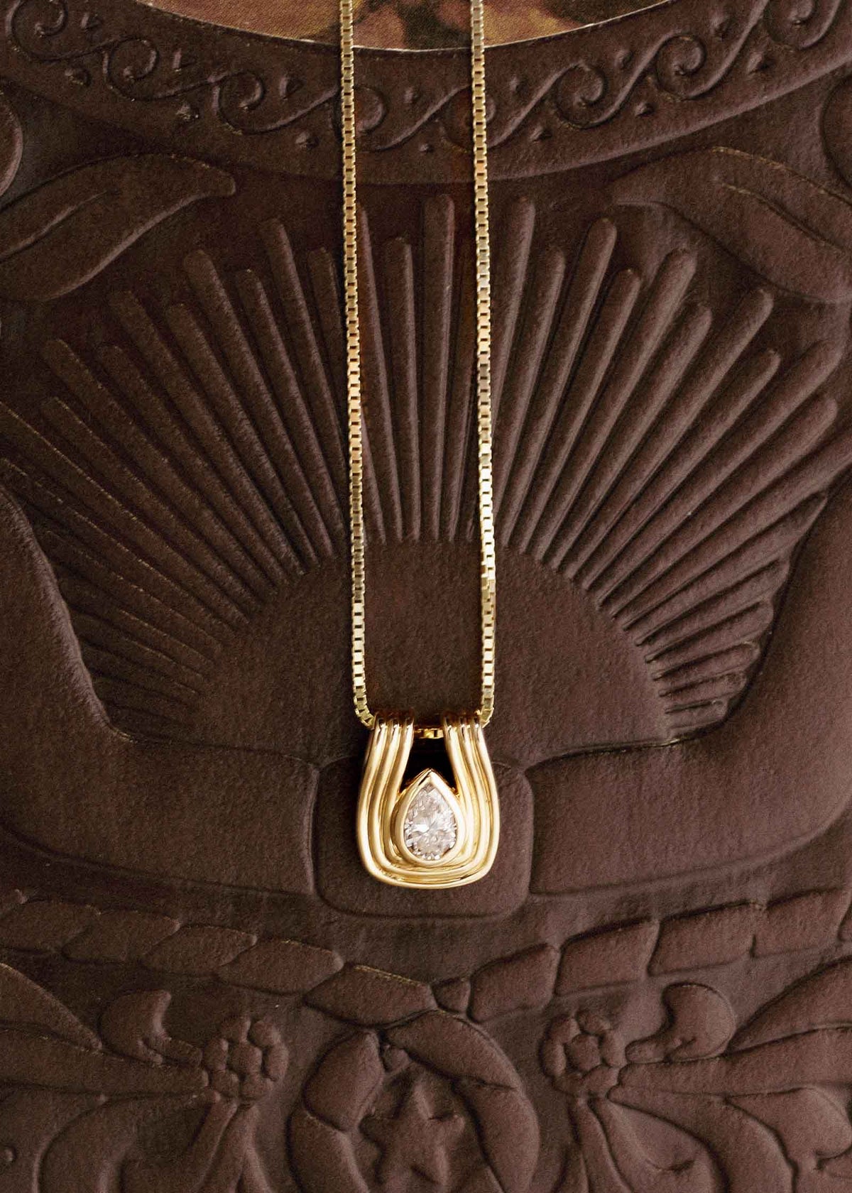 A Kinn Studio 14k Gold Alyssa Pear Diamond Pendant on a Box Chain laying against embossed leather