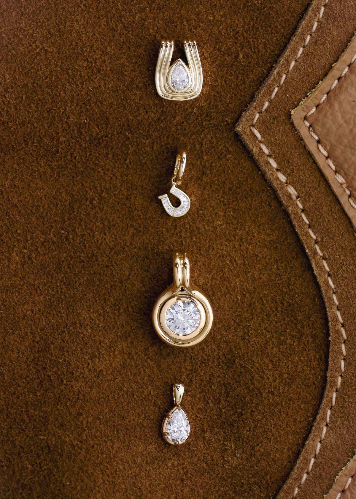 A Kinn Studio 14k Gold Jody Round Diamond Pendant, Clara Pear Diamond Pendant, Alyssa Diamond Pendant, and Mini Horseshoe Pendant laying flat against a brown suede background
