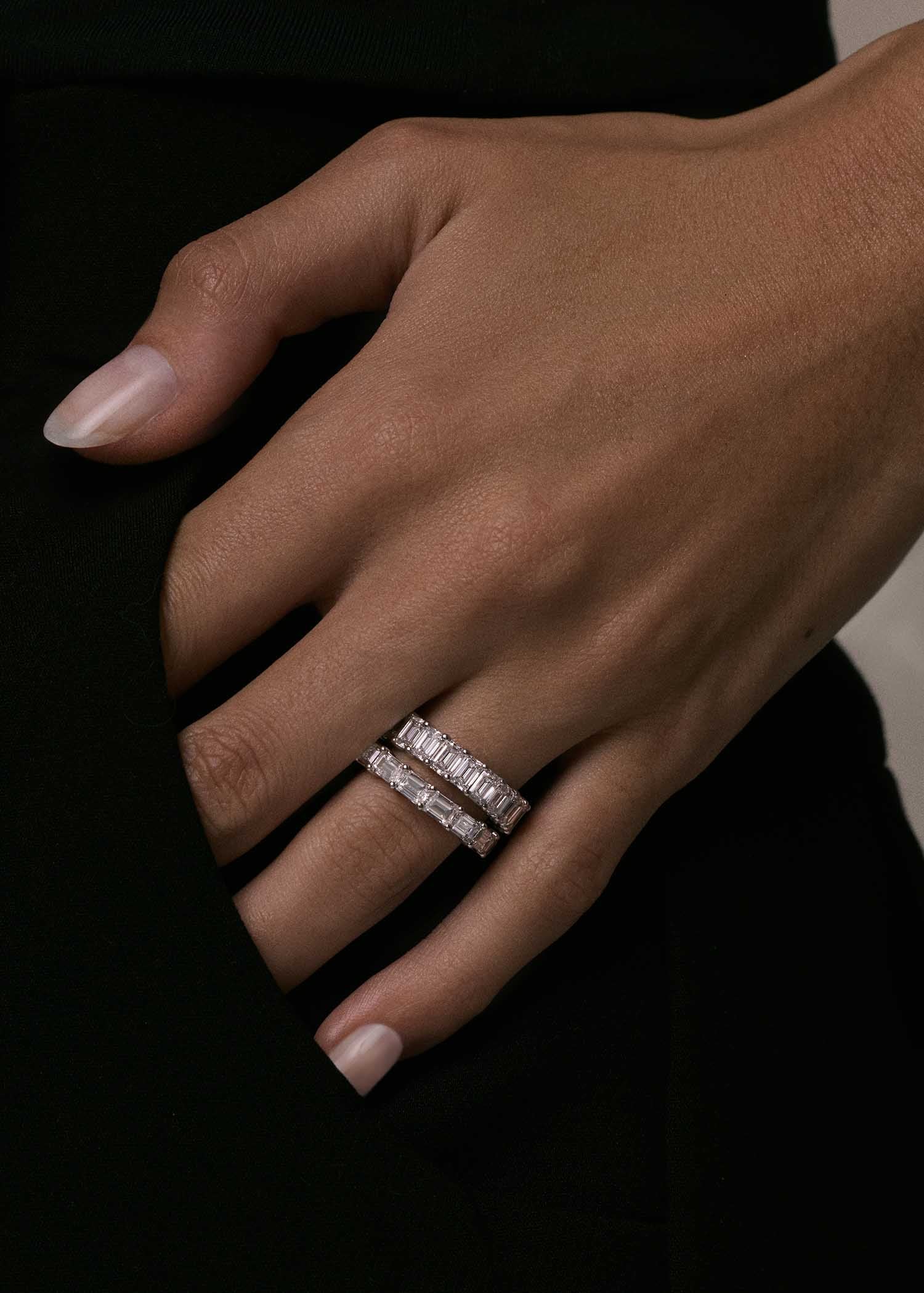 A close up of a woman's hand wearing a Kinn Studio 14k Gold Blaire Emerald Cut Diamond Eternity Band Ring in white gold stacked with a Jackie Emerald Diamond Eternity Band Ring
