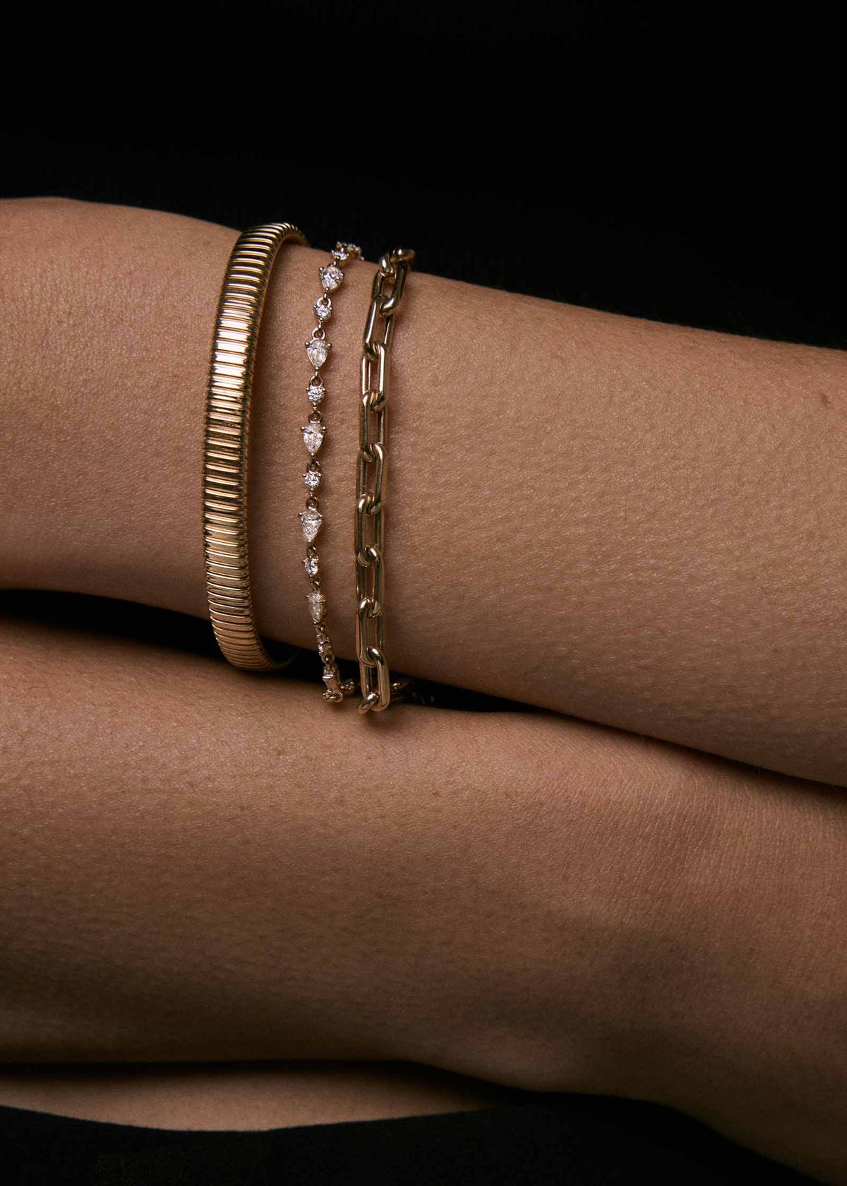 A close-up of a person's wrist wearing a Kinn 14k Gold Celina Flat Tubogas Bracelet stacked with a Mia Link Chain Bracelet and a Juliette Diamond Row Bracelet