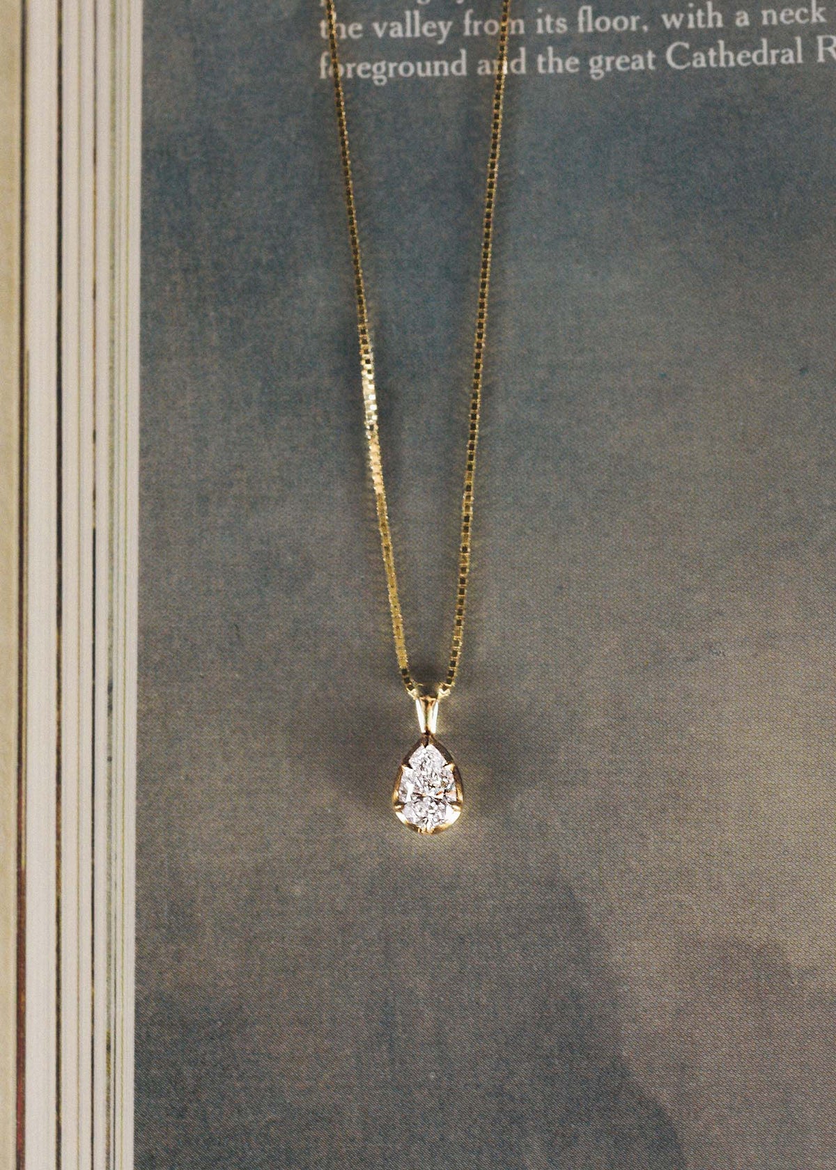 A Kinn 14k Gold Clara Pear Diamond Pendant on a box chain laying on a book
